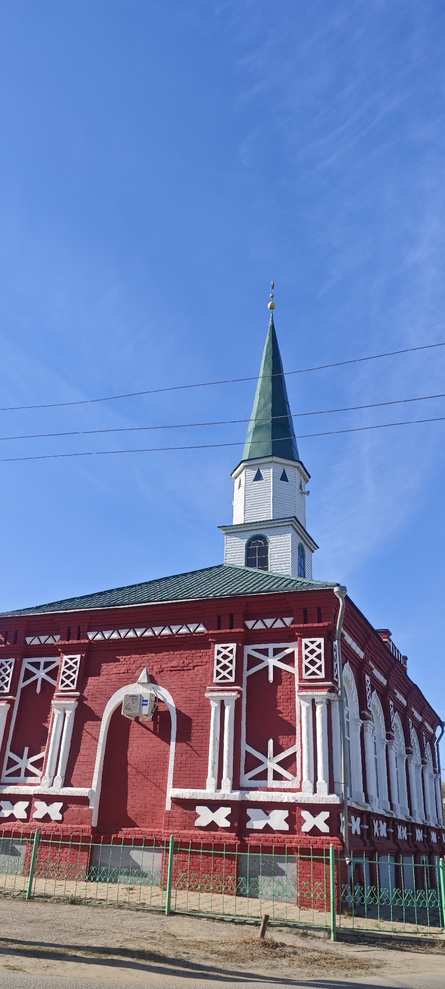 The image shows the First Cathedral Mosque of Khvalynsk, Russia, on a sunny day. The mosque is a red brick building with white decorative elements and a green roof. A tall, slender minaret with a green conical spire rises from the roof. A green metal fence surrounds the building. Power lines are visible against the clear blue sky. The ground in front of the building is unpaved. There are no people visible.