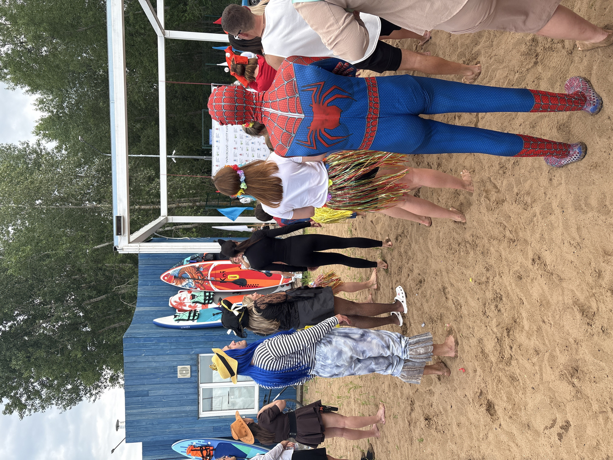 The image shows a group of people in costumes on a sandy beach. In the foreground, a person in a Spiderman costume is facing away from the camera, and a person in a colorful grass skirt is standing next to them. Further back, a person in a black cat costume, a person dressed as a pirate, and another in a striped shirt and tie-dye pants are visible. There are several paddleboards leaning against a blue wooden building in the background, suggesting the event might be related to water sports. Trees and a cloudy sky are also visible. The scene appears to be outdoors during the daytime. Some text is visible on a sign on the building, but it is mostly illegible. The atmosphere seems festive and playful, indicated by the costumes and the casual setting.