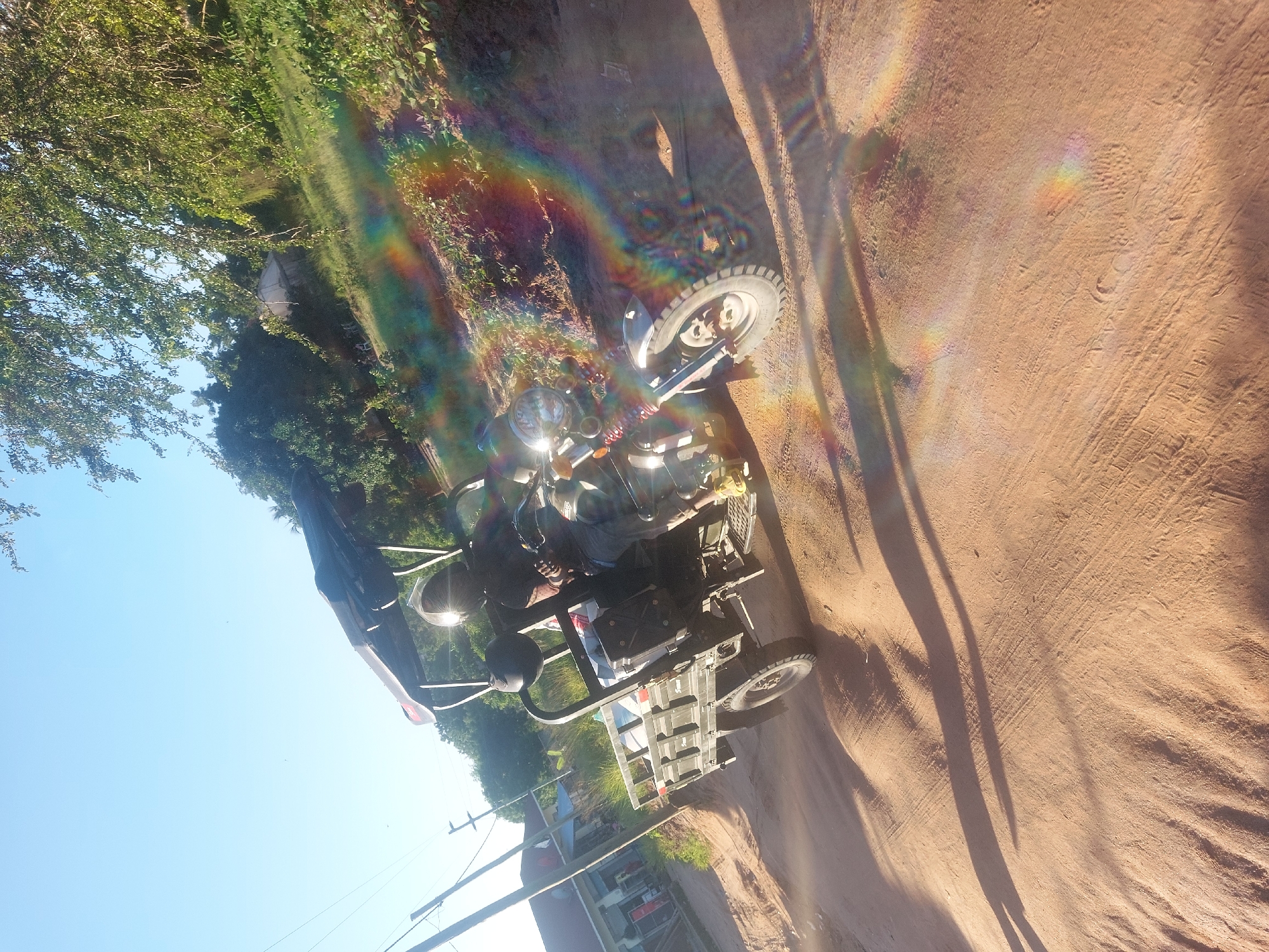 The image shows a three-wheeled vehicle on a dirt road. The vehicle appears to be a modified motorcycle with a cargo bed behind the driver's seat. The driver is wearing a helmet. The road is unpaved and sandy. There are trees and vegetation visible in the background. The location appears to be a rural or semi-rural area. Specific location information in Dar es Salaam, Tanzania, is not discernible from the image.