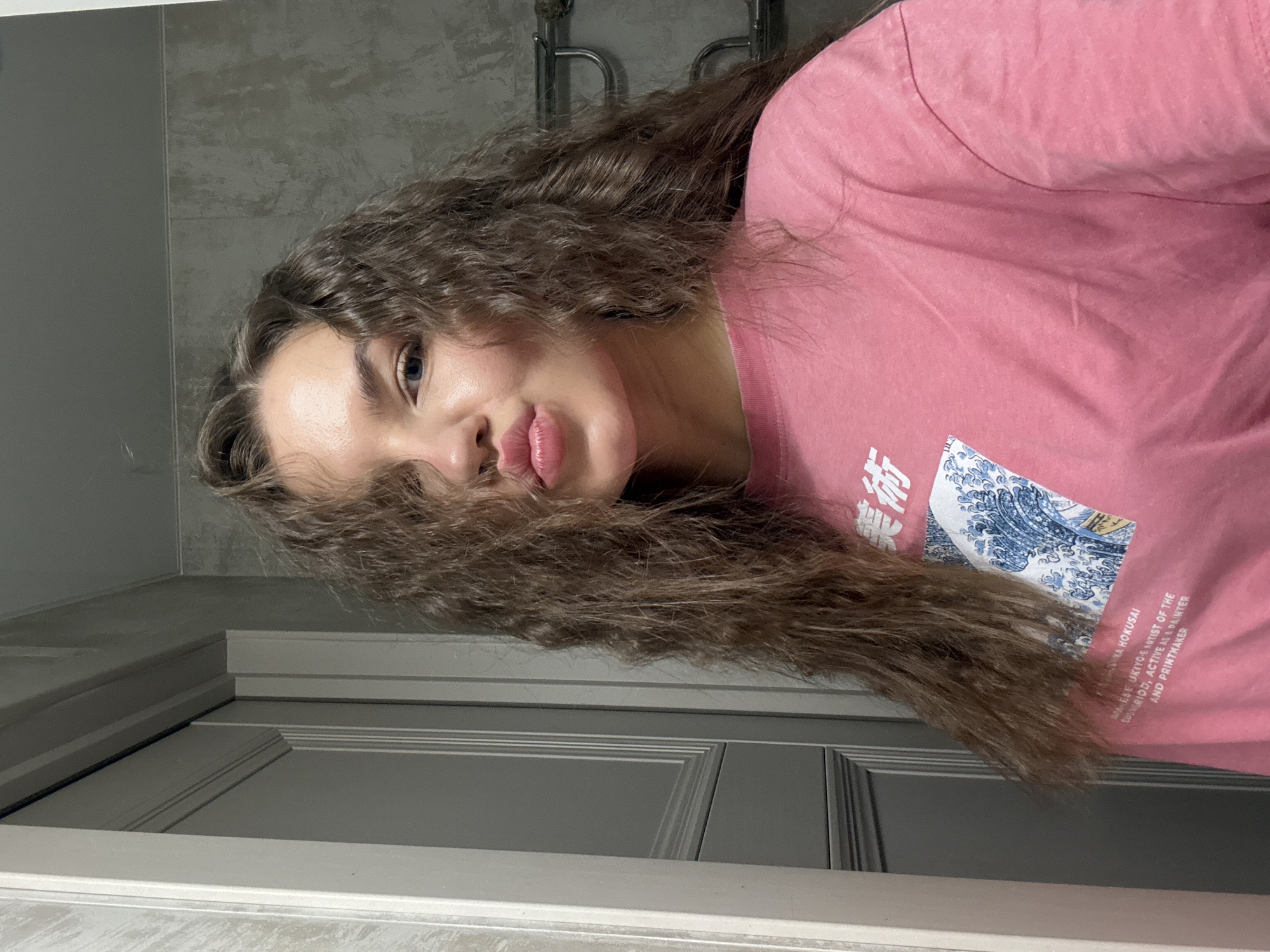 The image shows a woman with long, curly brown hair partially obscuring her face. She is wearing a pink t-shirt with Japanese text and a wave design printed on it. The background includes a gray tiled wall, a metal towel rack, and a gray door. The location is likely inside a bathroom or similar interior space. The city of Moscow, Russia, is not identifiable from the image.