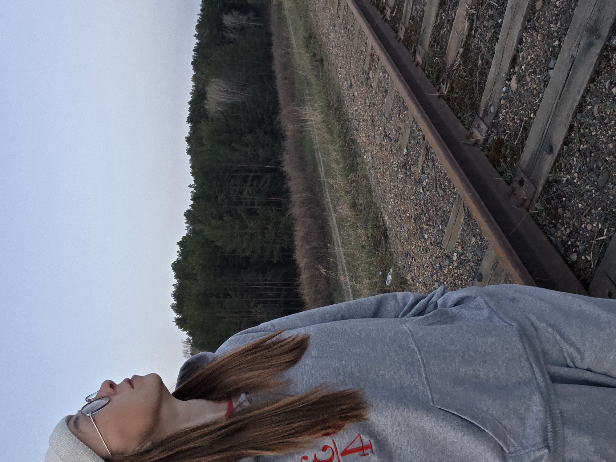 A woman in a gray sweatshirt and a white beanie lies on a railway track. She is wearing glasses and has long, reddish-brown hair. The track runs through a landscape of trees and grass under a light gray sky. The location is near Chaikovsky, Russia.