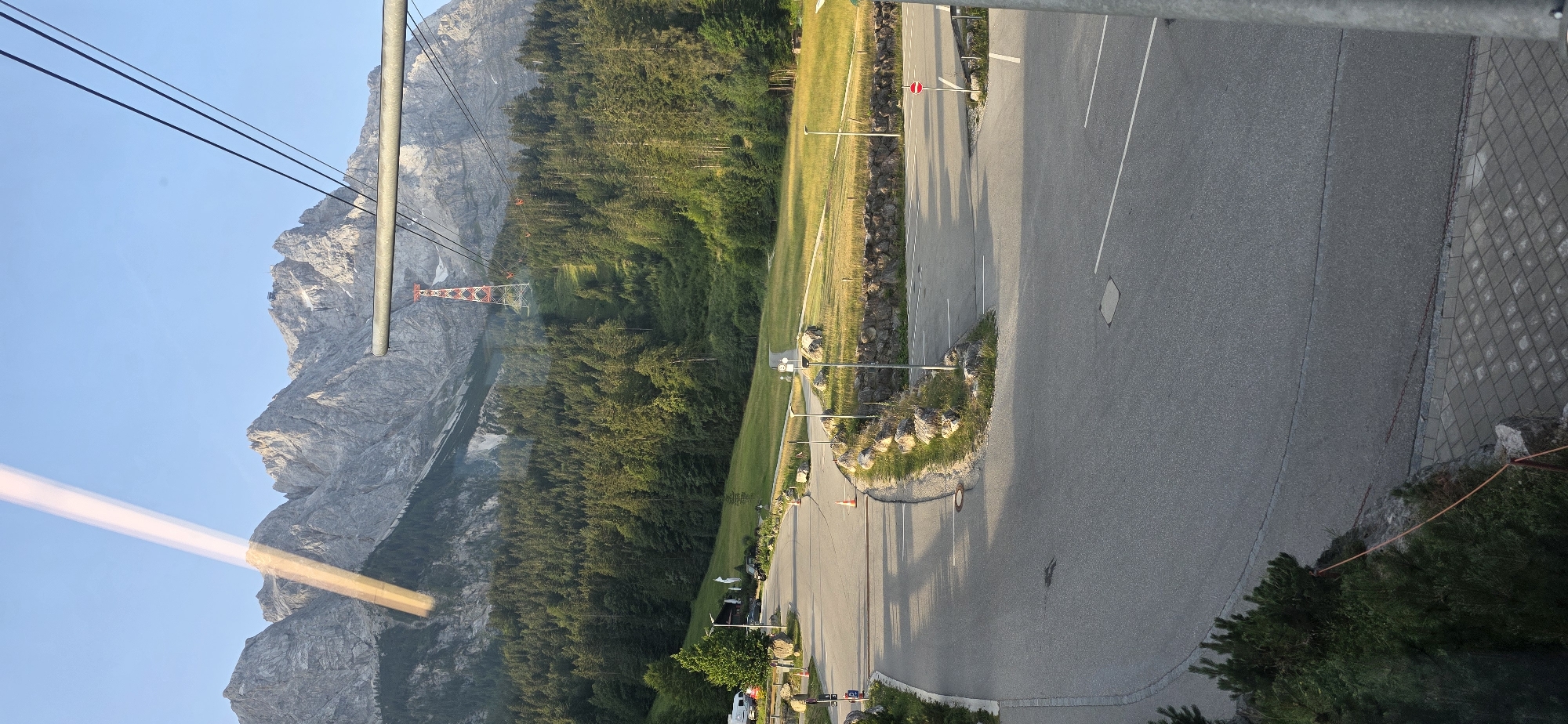 The image depicts a scenic mountain landscape in Grainau, Germany, likely taken from a cable car. In the foreground, a paved road curves through a parking area with white parking lines. Several large rocks are scattered along the roadside. Beyond the road, a grassy field slopes gently upwards, bordered by a dense forest of evergreen trees. In the background, a towering mountain range dominates the skyline, with rugged peaks reaching towards a clear blue sky. A red and white transmission tower is visible on the mountainside, with cables extending from it towards the foreground, suggesting the presence of a cable car system. The scene is bathed in the warm glow of sunlight, indicating it is either early morning or late afternoon.