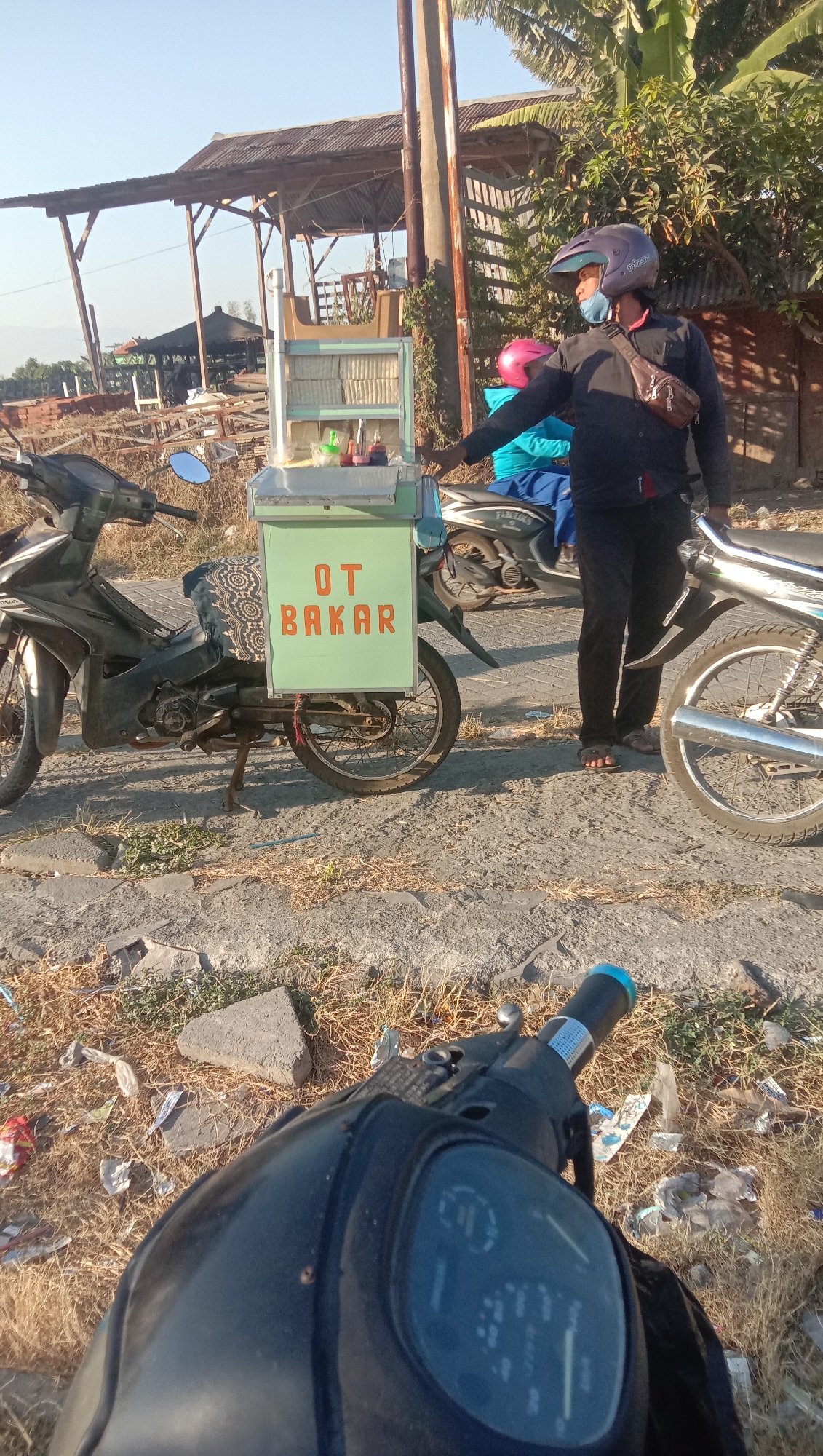 In Curah Dukuh, Indonesia, on a sunny day, a man wearing a dark long-sleeved shirt, dark pants, sandals, and a blue helmet is standing next to a food cart attached to a motorcycle. The cart is green with "OT BAKAR" written in orange letters on the side. There are bottles of condiments and food items displayed inside the cart. A woman wearing a pink helmet and a blue shirt is seated on a motorcycle behind the food cart. Another motorcycle is parked to the left of the food cart. The ground is made of gravel and dirt, with dry grass and scattered litter. In the background, there are structures with corrugated metal roofs and trees. The scene appears to be a roadside food stall.