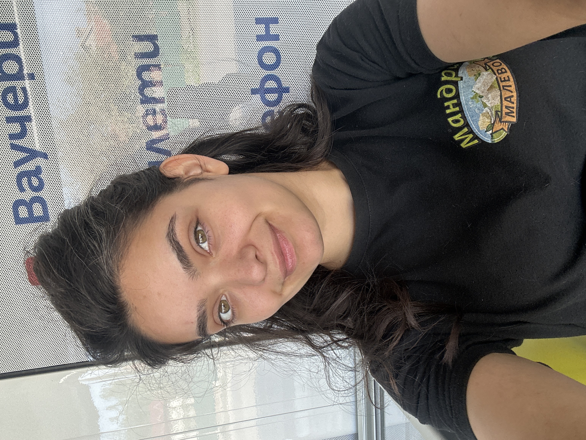 A young woman with dark hair and green eyes is taking a selfie. She is wearing a black t-shirt with a circular emblem featuring text and images. The background appears to be a bus stop or similar public transport area, with perforated metal signage displaying text in Cyrillic script. One of the visible words is "Malevo," indicating the location in Bulgaria. The lighting suggests it is daytime.