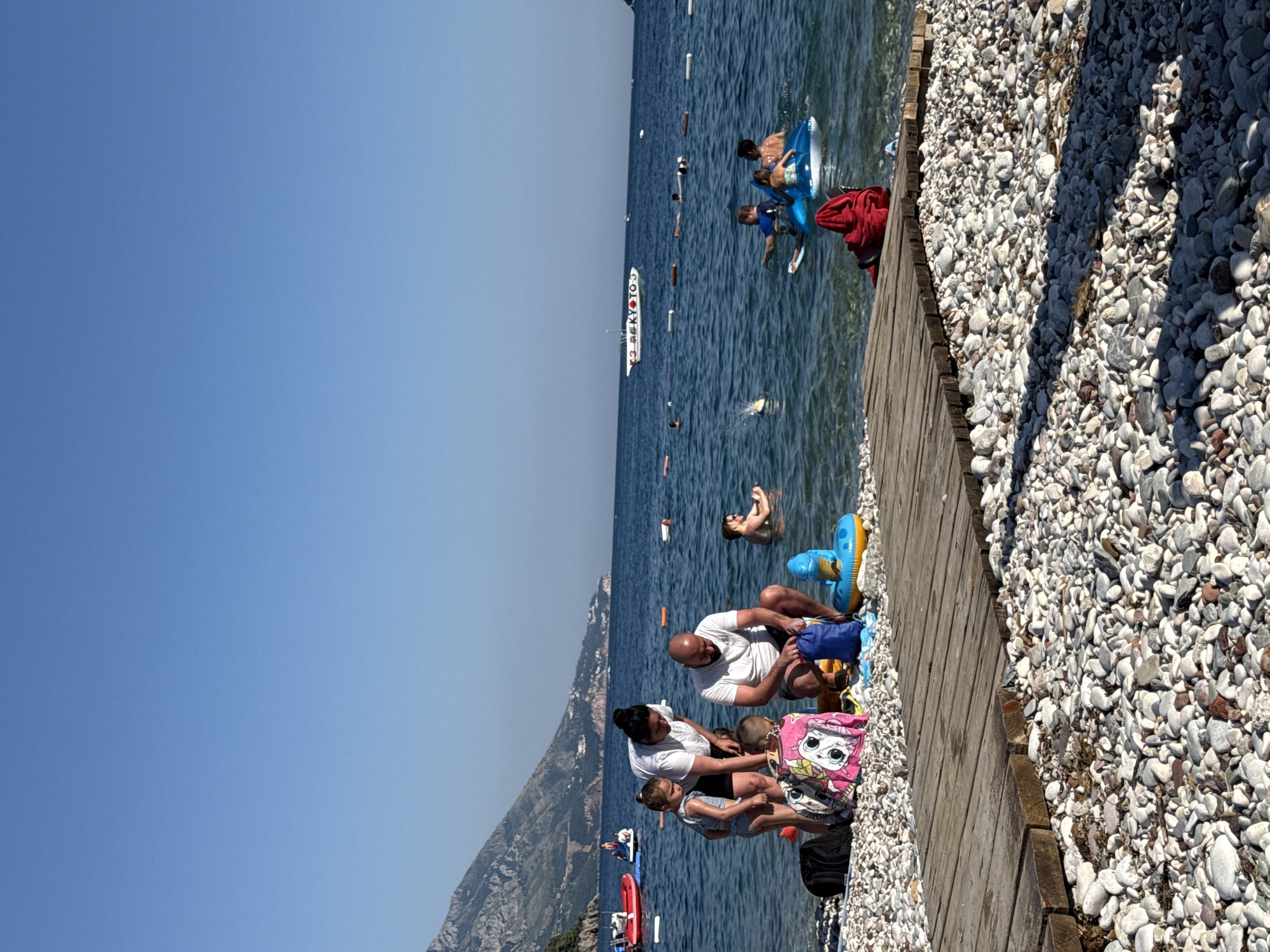 In Budva, Montenegro, on a sunny day at a pebble beach, a family is gathered near the water's edge. A man and a woman, presumably parents, are interacting with two young children. The woman is bent over, attending to one of the children, a girl in a swimsuit. The man is kneeling and appears to be packing or organizing a blue bag. Beside them are beach accessories including towels and a blue inflatable toy. In the shallow water, several other people are visible. A man is sitting in the water, and further out, two individuals, likely older children or teenagers, are on a blue inflatable. A boat with the text "B" and "TO" is in the background, along with more buoys marking a swimming area. The background features a mountain range and a clear blue sky. A wooden walkway made of planks is in the foreground, laid across the pebbles.