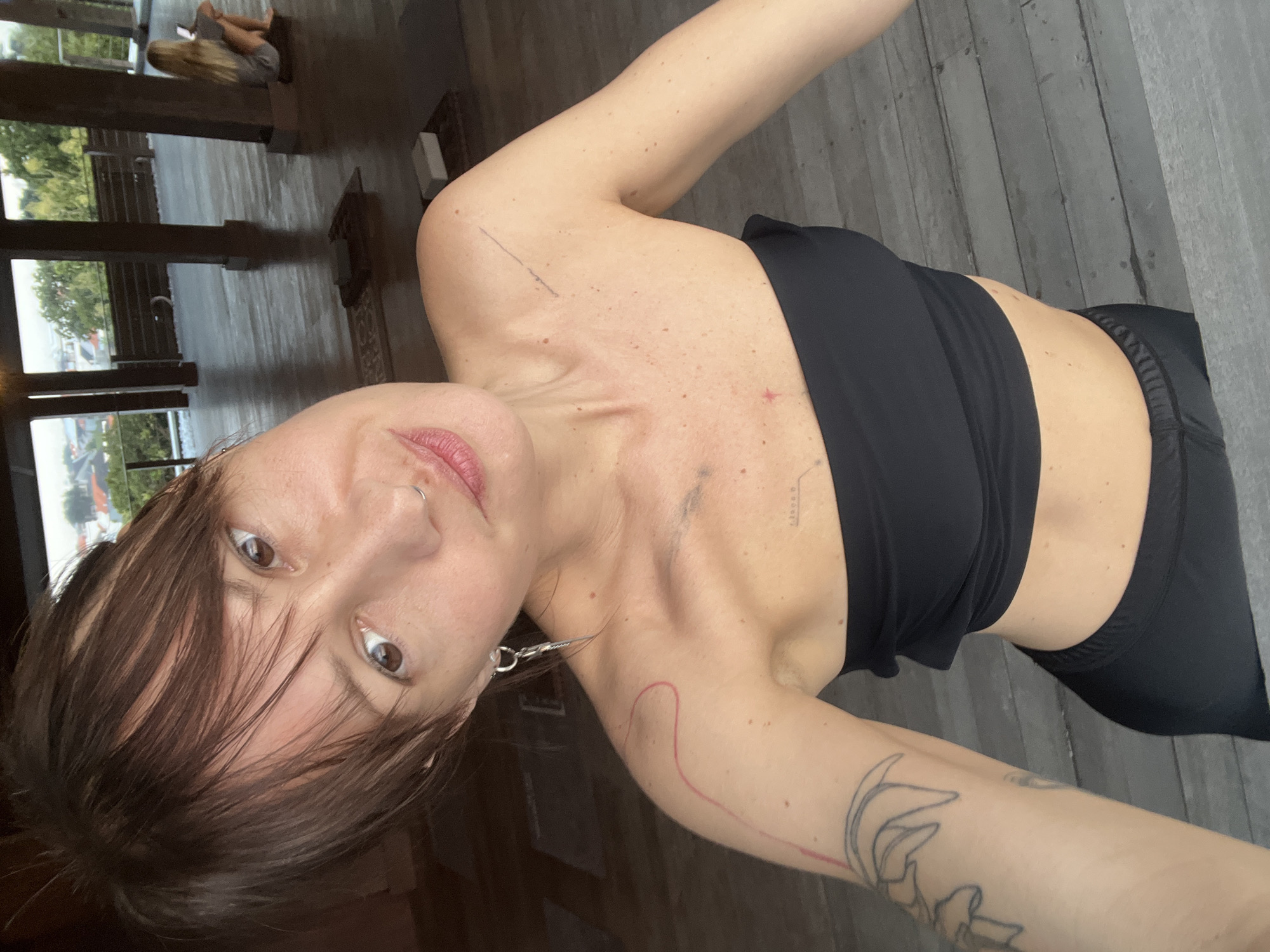 A woman with short brown hair, wearing a black sports bra and black leggings, is posing for a selfie. She has a nose ring and a silver earring hanging from her left ear. She has several tattoos on her body. She is standing on a wooden deck. In the background is a building with large windows looking out over a body of water. In the far distance, there is another person sitting on the deck, looking at a tablet. The location appears to be a building in Mengwi, Indonesia.