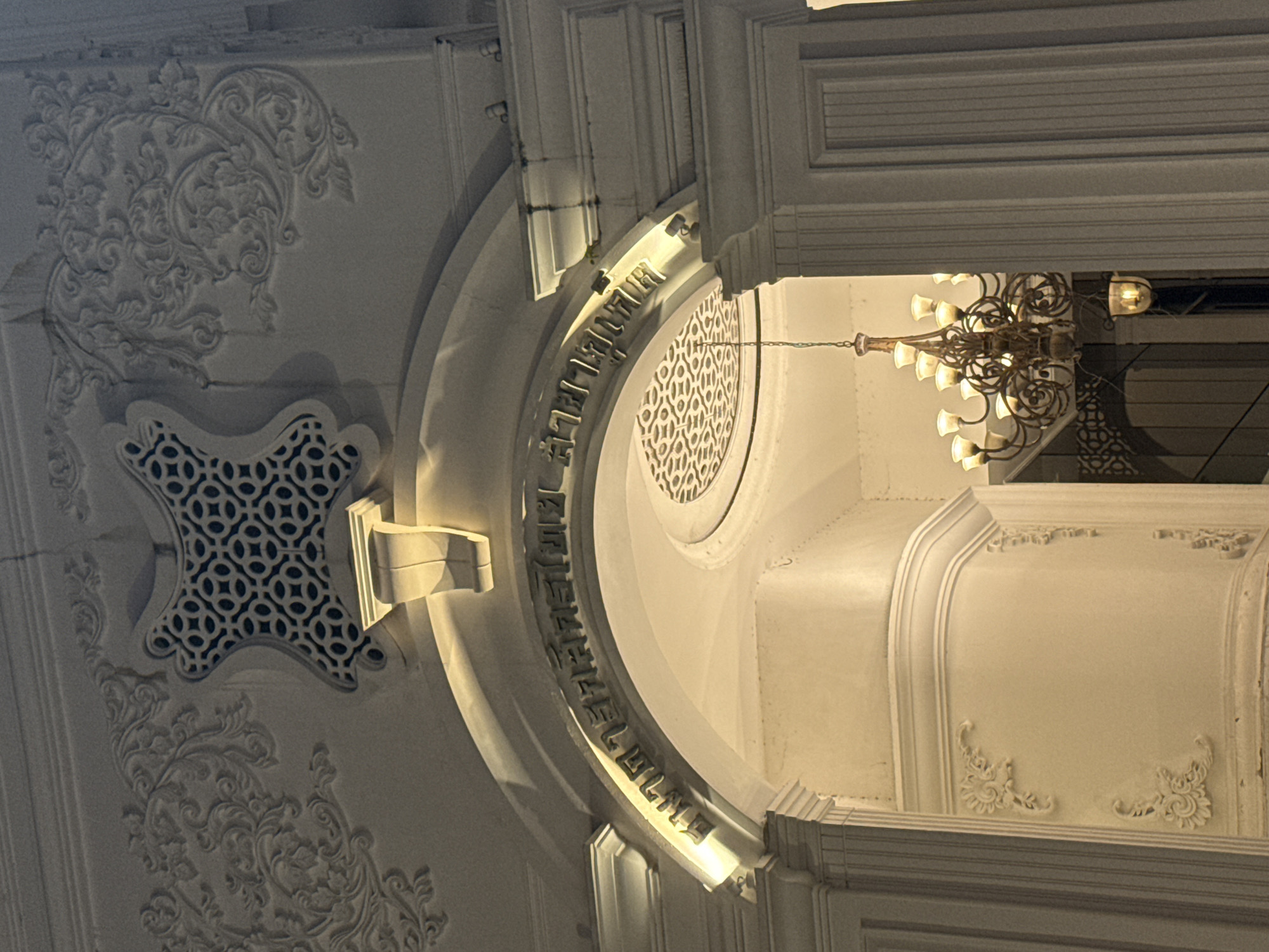 The image shows the entrance to the Phuket Thai Hua Museum in Phuket City Municipality, Thailand, at night. The building features ornate architectural details, including intricate carvings and a decorative grille above an archway. Golden text in Thai script is inscribed along the arch. A chandelier is visible inside the building, illuminated by warm light. The scene is lit by external lights highlighting the building's facade.