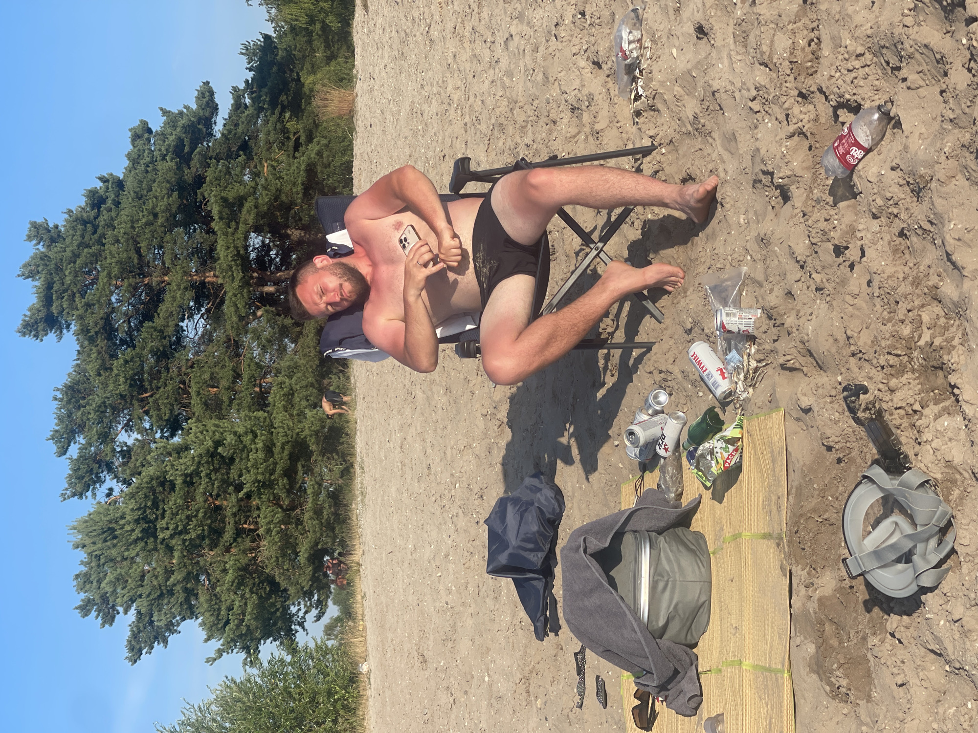 A man is sitting in a folding chair on a sandy beach. He is wearing black swim trunks and is shirtless. He is holding a smartphone in his hand. Several empty cans and plastic bottles are scattered on the sand around him, along with a towel draped over a cooler, a pair of sunglasses, and a pair of flip-flops. In the background, there are tall pine trees and a clear blue sky. Other people can be seen in the distance among the trees.