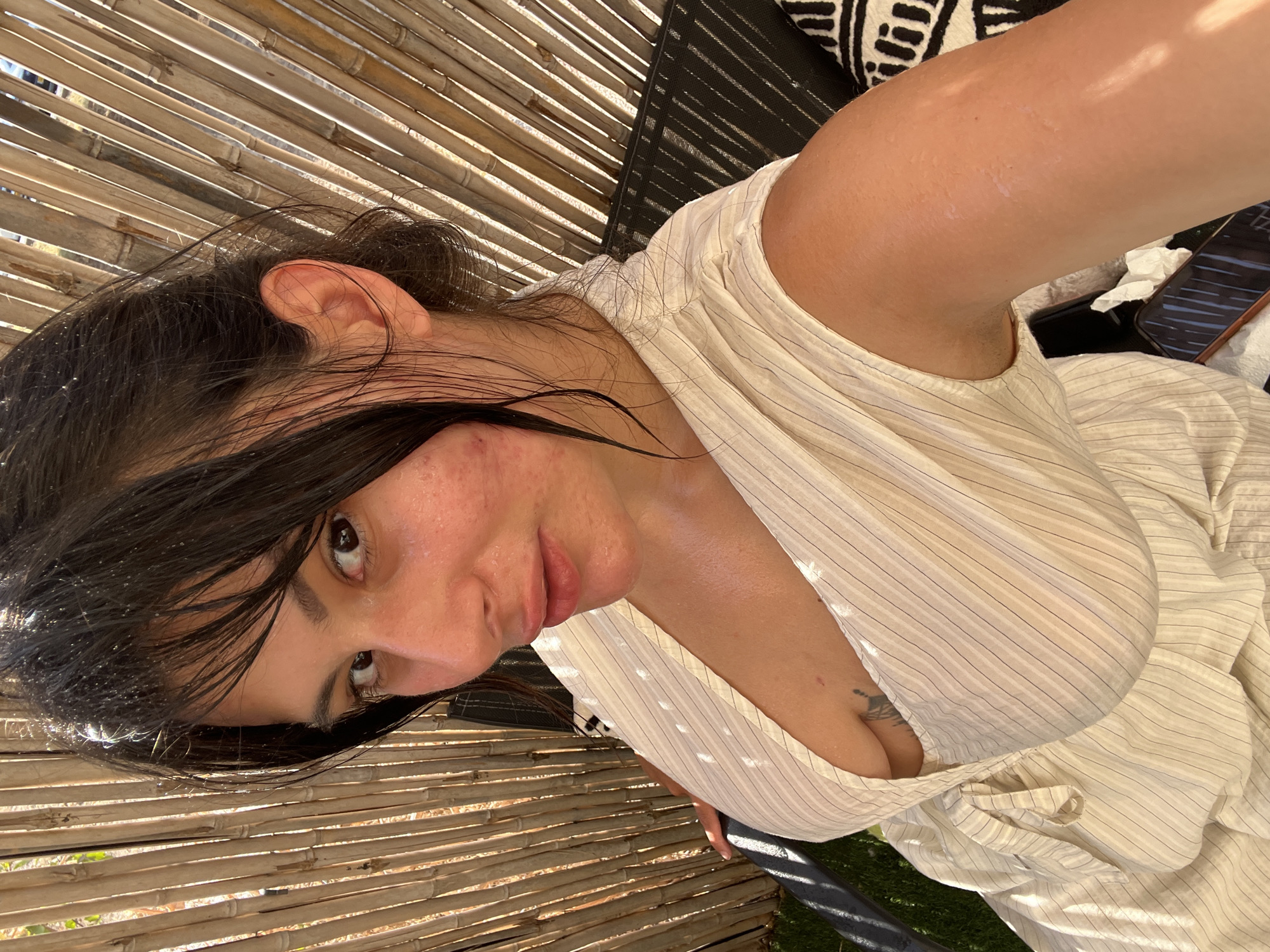 A medium shot captures a woman with dark hair pulled back, some strands falling loose around her face and neck. She is outdoors, seated on a dark-colored chair with a patterned cushion, in front of a bamboo screen. Sunlight dapples through the screen, casting shadows on her face and light-colored, striped dress, which is open at the front, revealing her chest. Her skin appears to be glistening, possibly from sweat or a lotion. A tattoo is visible on her chest. The background suggests a natural setting with grass visible to the left. The image was taken in Kiryat Yam, Israel.