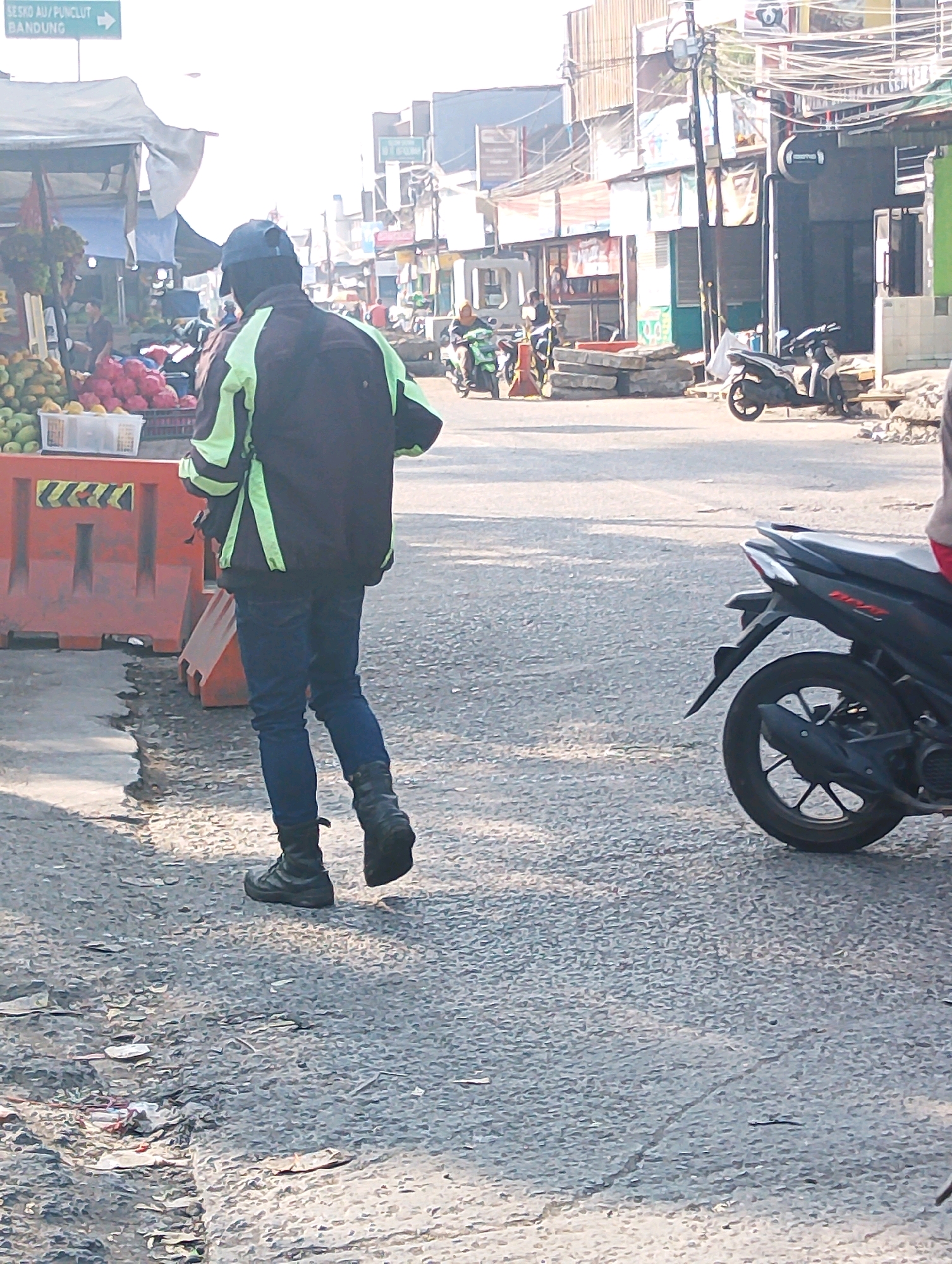 The image depicts a street scene in Lembang, Indonesia. A person wearing a dark jacket with light green accents and blue jeans walks away from the camera on a paved road. They are wearing dark boots and a dark baseball cap. To the left of the person, an orange traffic barrier is visible, with a market stall behind it displaying fruits and other produce under a white canopy. In the background, various buildings and shops line the street, with power lines overhead. A parked black motorcycle is visible on the right side of the road. The overall environment appears to be a busy street market area.