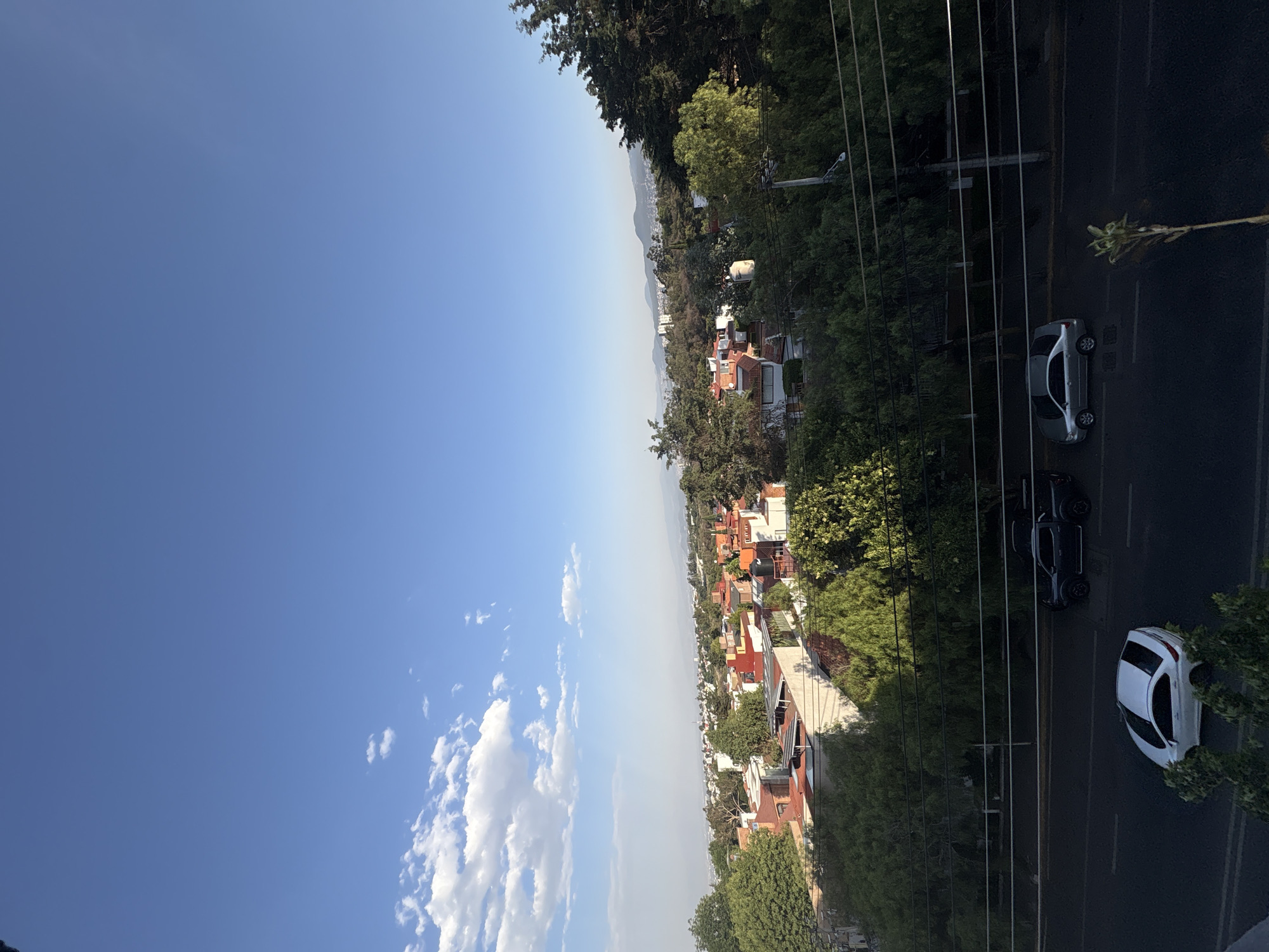 The image shows a cityscape, likely within Mexico City, Mexico, characterized by a clear blue sky above and a dense urban area below. The urban environment is comprised of numerous buildings, with a mix of red-tiled roofs and modern structures, interspersed with lush green trees, creating a layered urban landscape. A road runs through the scene, and vehicles, including a white car, a black truck, and a silver sedan, are visible on it. Utility lines are seen crisscrossing above the road and trees. The overall atmosphere is bright and sunny.