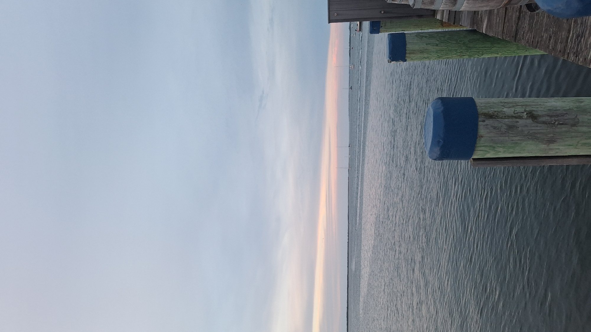 The image depicts a serene waterscape with a view of the ocean and sky. In the foreground, wooden pilings with blue caps are visible along a pier. The water is calm, with subtle ripples on its surface. In the distance, several sailboats are dotted across the horizon, adding a sense of tranquility to the scene. The sky is a pale blue, with soft, wispy clouds that hint at the possibility of dawn or dusk. The overall atmosphere of the image is peaceful and inviting, suggesting a quiet moment by the sea.