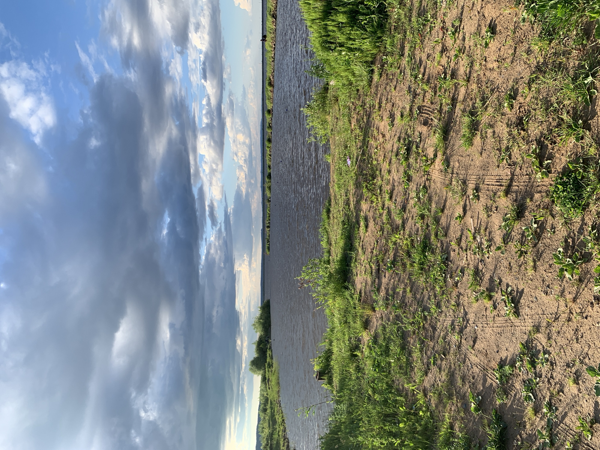 A wide shot captures a view of a lake under a dramatic sky in Chaikovsky, Russia. The foreground is a sandy area with sparse green vegetation and tire tracks. The midground features the calm, dark water of the lake, bordered by lush green grass and small bushes. In the distance, a line of trees and vegetation stretches across the horizon. The sky is a mix of heavy, dark clouds and patches of bright blue, with sunlight breaking through in places. Two figures are visible on the far bank of the lake. The overall scene suggests a natural landscape under changing weather conditions.