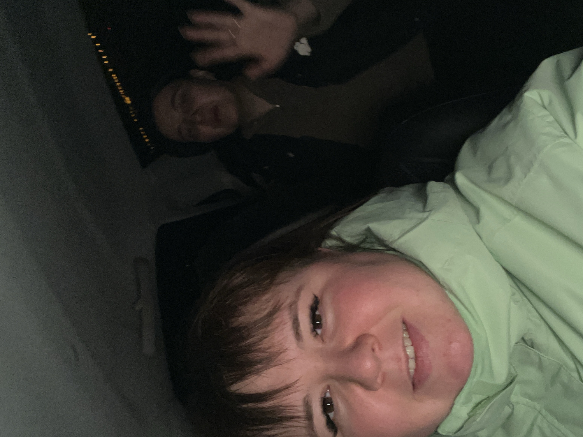 A rotated selfie shows two people in a dimly lit environment. The person in the foreground is smiling, facing the camera, and wearing a light green jacket. The second person is behind them, partially visible, with a hand raised. A line of lights is visible in the background through a window. Based on the prompt, the location is implied to be Chaikovsky, Russia.