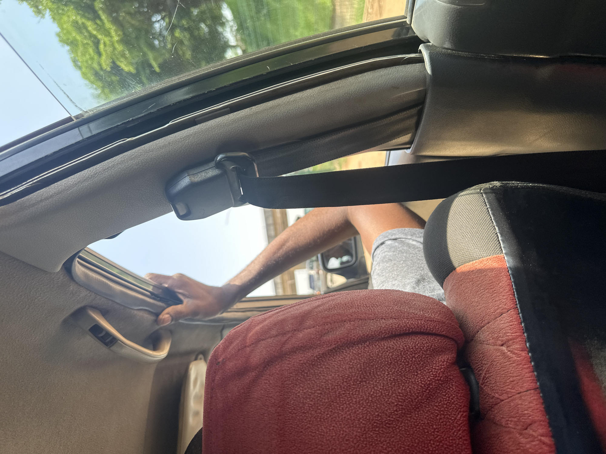 The image depicts the interior of a car, looking out through the passenger side window. In the foreground, a portion of a red car seat is visible, with the headrest to the left and the main seat to the right. A black seatbelt mechanism is prominent in the center, with its strap hanging down. A person's arm, resting on the car's doorframe and reaching towards the window handle, is visible in the mid-ground. The window is partially open, revealing an outdoor environment with trees and a bright sky. In the distance, a road with vehicles and an overhead bridge can be seen. The scene is consistent with a journey within the city of Abuja, Nigeria, given the typical vehicle interiors and outdoor scenery found in the region.