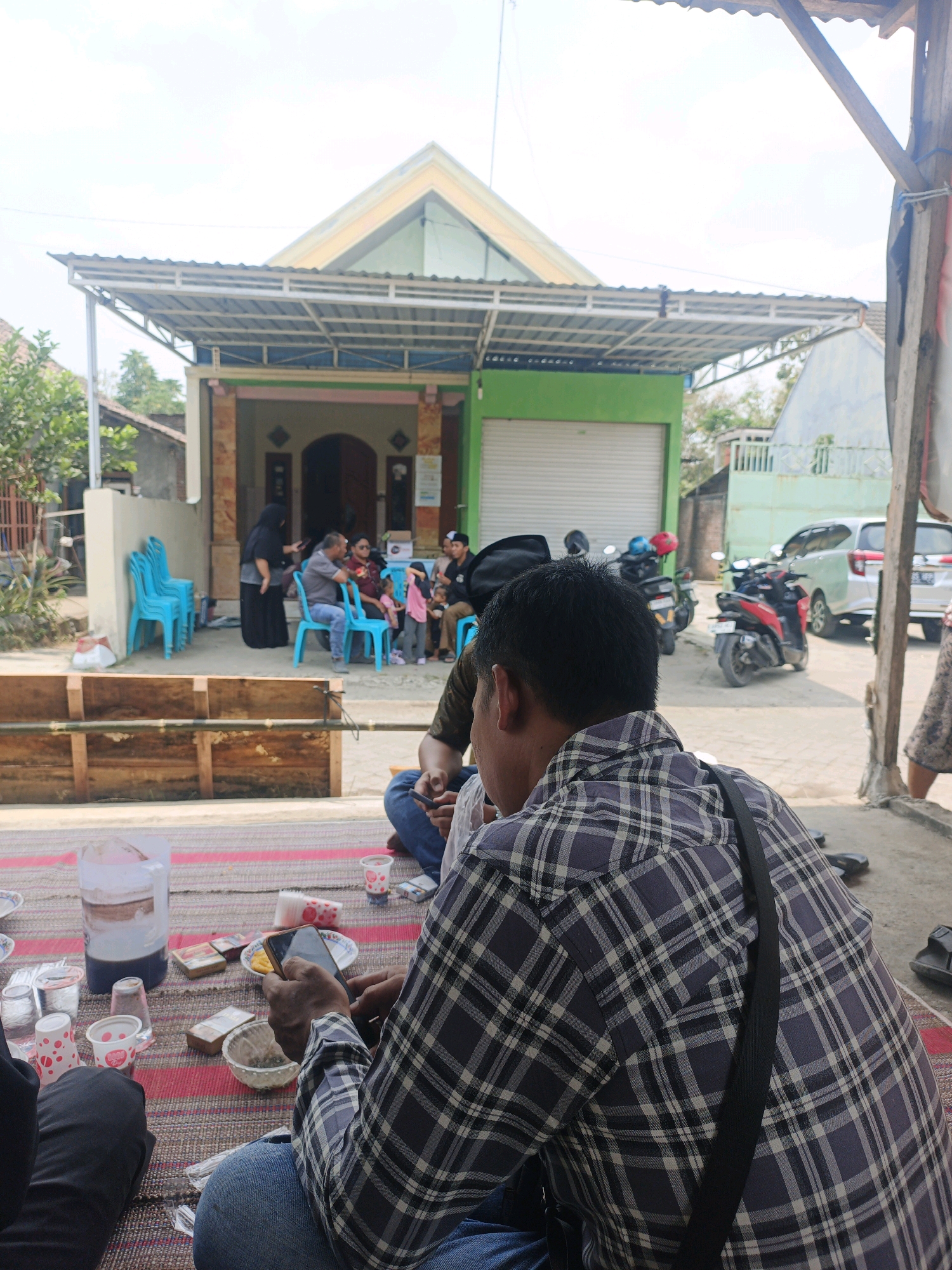 In Mojoduwur, Indonesia, a group of people are gathered outdoors in front of a building with a green garage-style door. The scene appears to be a casual social gathering or meeting. In the foreground, a person wearing a plaid shirt is looking at their phone while sitting on a mat. Several items are spread on the mat, including a large jug of liquid, cups, cigarette packs, and a small bowl. In the middle ground, several individuals are seated on blue chairs, with some appearing to be in conversation. Behind them, the building is visible, with a covered outdoor area. Several motorcycles are parked near the building, and a silver car is parked further in the background. The environment suggests a warm, sunny day.