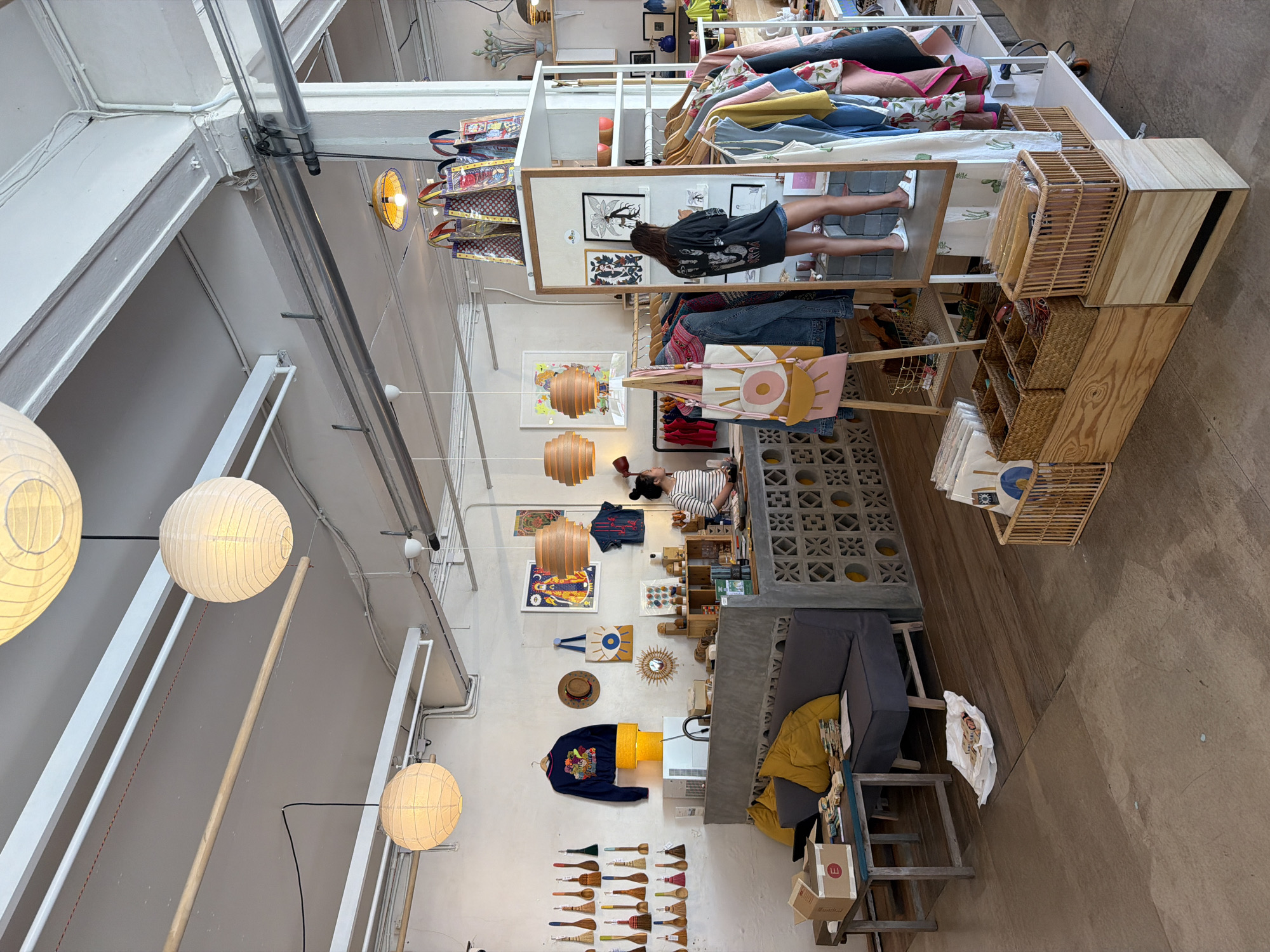 This is an indoor scene of a retail shop, likely a boutique or gift shop, located in Chiang Mai City Municipality, Thailand. The shop has a rustic and bohemian aesthetic with exposed concrete, wooden elements, and minimalist decor.
In the foreground, a dark wooden floor transitions to a lighter-colored stone floor. A grey couch with yellow pillows is placed against a concrete counter. On the left, a wooden table holds a cardboard box, a laptop, and stacks of books or papers.
The counter area features a display of brushes and spatulas on the wall. Behind the counter, two individuals are visible. One person, wearing a striped shirt, appears to be an employee, possibly attending to the counter. Another person, a woman with dark hair, is standing in front of a full-length mirror, examining clothing on a rack. She is wearing a dark oversized t-shirt and shorts, with white sandals.
Various items are displayed throughout the shop, including clothing on racks, handbags hanging from displays, artwork on the walls, and decorative objects. Several round, paper lanterns are suspended from the ceiling, providing diffused lighting. The overall atmosphere suggests a bright and airy retail space.