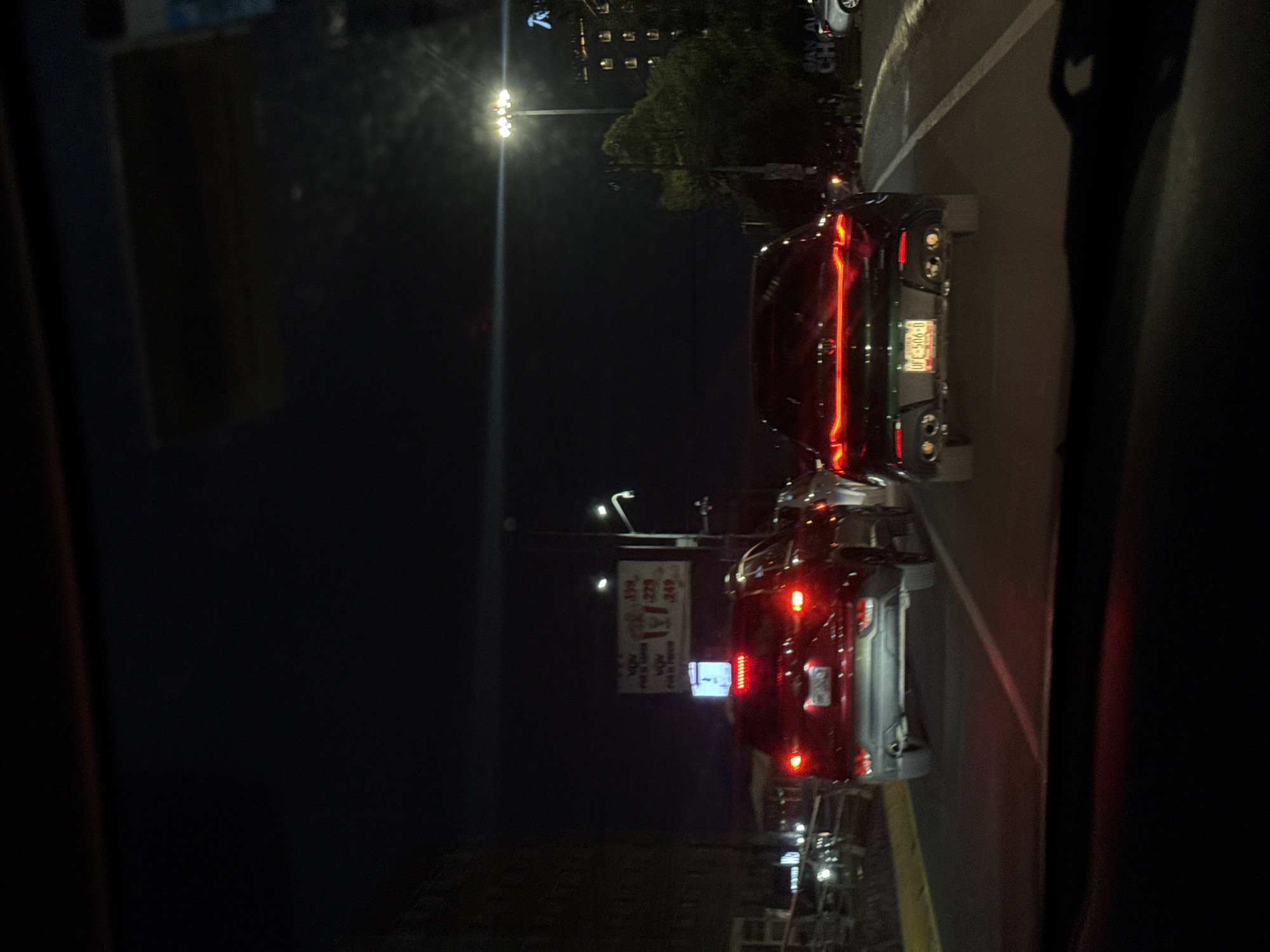 The image depicts a night scene in San Bernardino Tlaxcalancingo, Mexico. Two cars are visible, one directly behind the other. The leading car is black, with illuminated red tail lights and a license plate with the identifier "UFE-906-D". A sign advertising pizza is visible, with the prices "$399," "$229," and "$249." Street lights illuminate the area, and a building with a "R" logo is visible in the background. The image appears to be taken from inside a car. The immediate surroundings are dark, with the car's interior barely discernible.