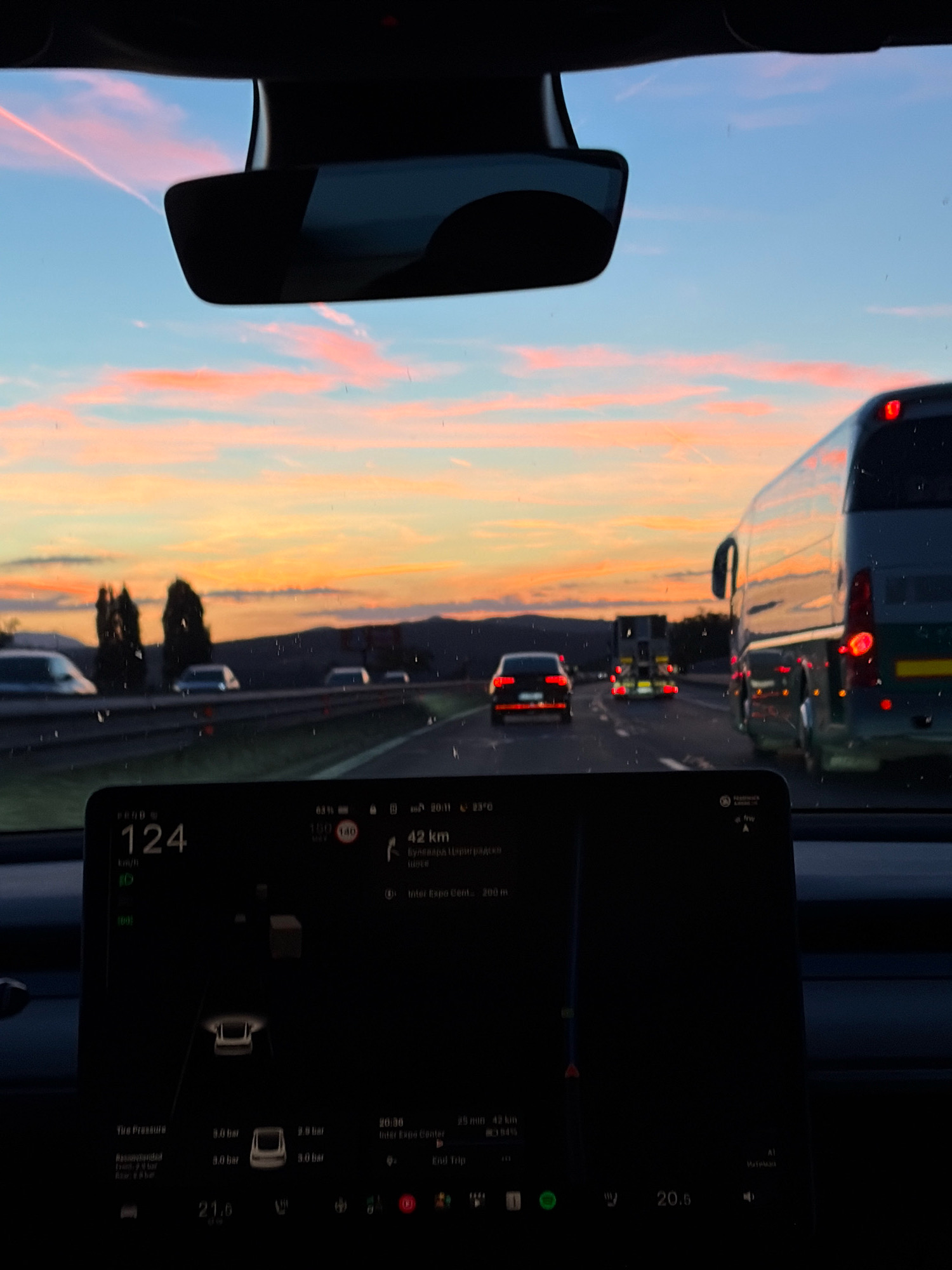 The image shows a view from inside a car, likely a Tesla, as indicated by the large touchscreen display in the foreground. The display shows vehicle speed at 124 km/h, navigation information with a destination 42 km away, and tire pressure readings. The car is on a highway at dusk, with a colorful sky displaying hues of orange, pink, and blue, suggesting either sunrise or sunset. Multiple cars and a large bus are visible on the highway ahead, illuminated by their headlights and taillights. The environment appears to be a highway with some trees and hills in the distance. The exact location is Unknown, Unknown.