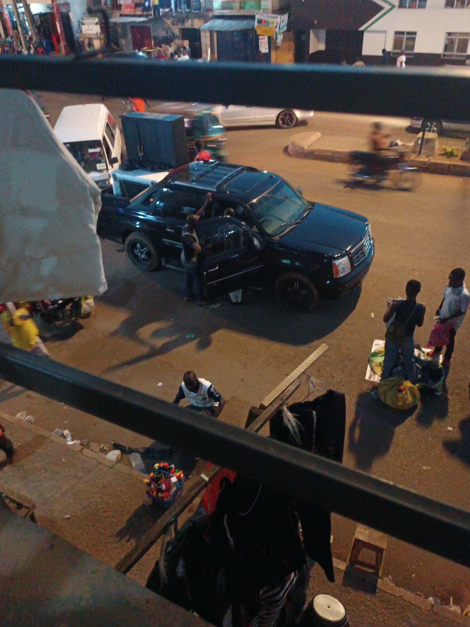 The image depicts a street scene at night in Unknown, Unknown. A black SUV is parked with its driver's side door open, and a man is standing near the open door. Several individuals are gathered nearby, appearing to be engaged in some sort of transaction or activity involving clothing or merchandise spread out on the ground. A white van is parked behind the SUV, and other vehicles and pedestrians are visible in the background. The scene is illuminated by streetlights and the lights of surrounding buildings. A railing in the foreground partially obscures the view.