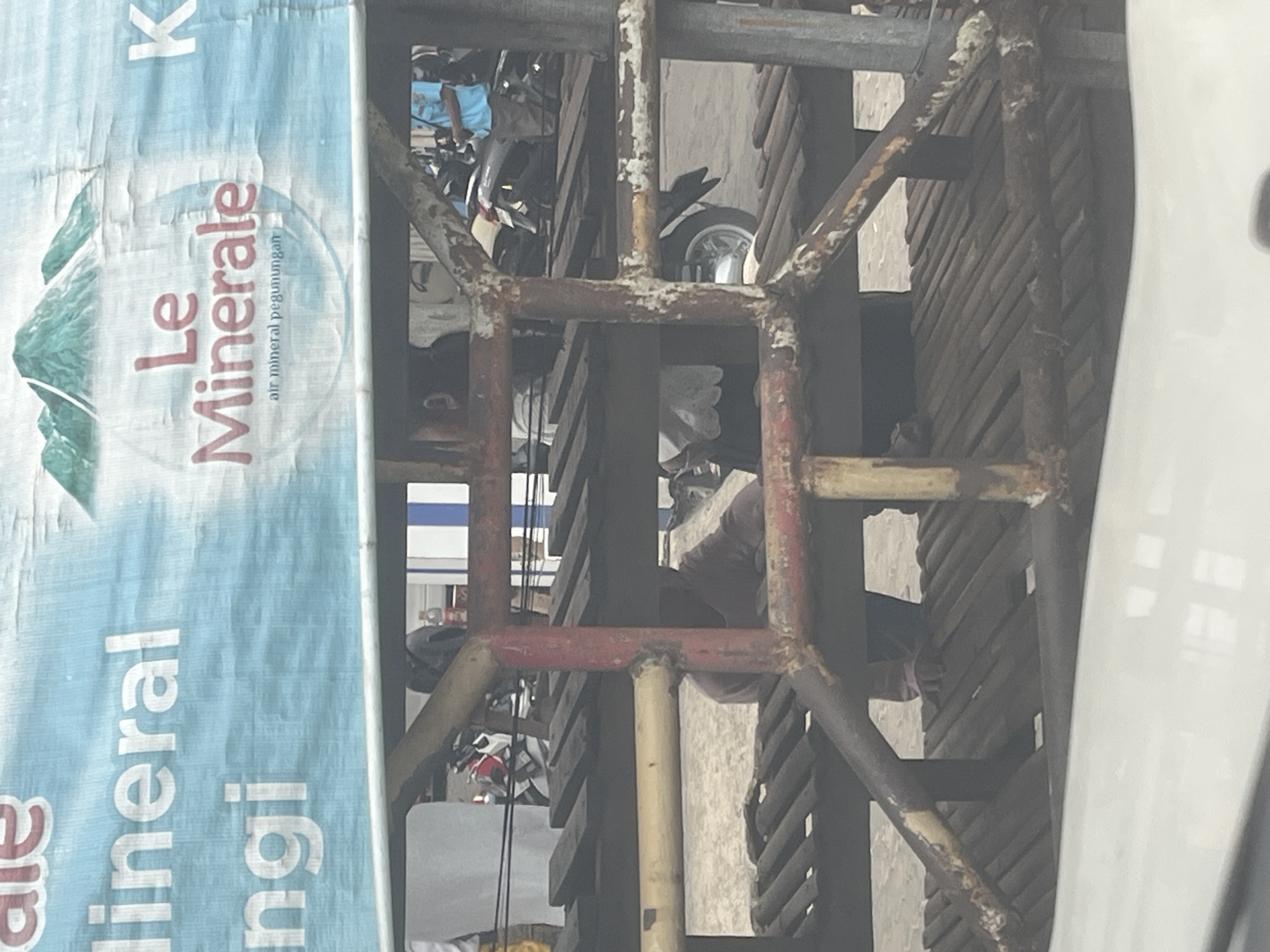 The image captures a street scene in Cilegon, Indonesia, dominated by a large, light blue banner advertising "Le Minerale" bottled water, featuring a mountain graphic. In the foreground, a rusty, metal structure, likely part of a temporary seating area or scaffolding, partially obscures the view. Beneath this structure, two individuals are visible, seated and facing away from the camera. One person, wearing a dark shirt, is hunched over, while another person in a light-colored top appears to be interacting with something in their lap. The background reveals more of the urban environment, with a few motorcycles and other people milling about. The overall impression is one of a typical street corner in a bustling Indonesian city.
