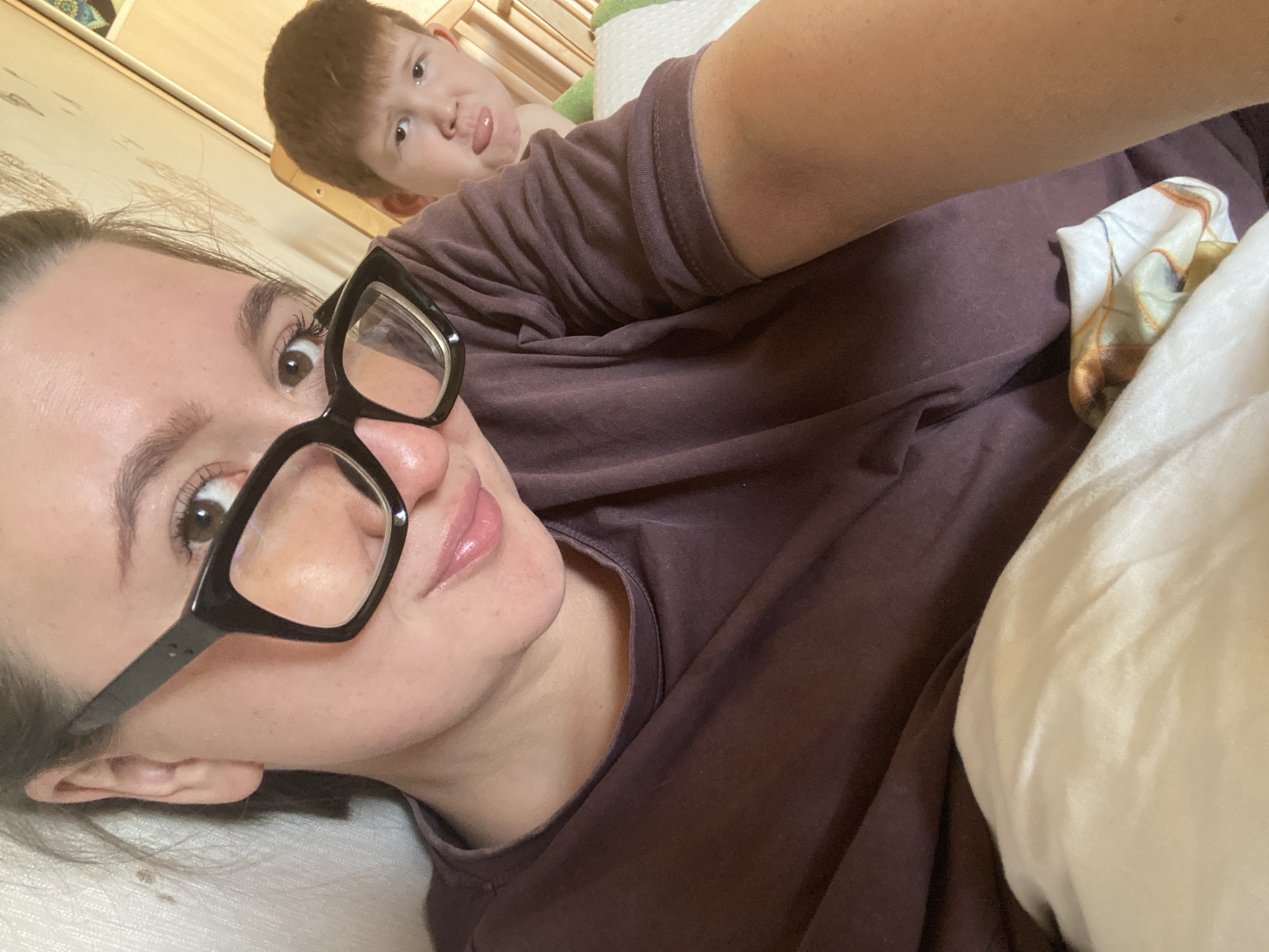 A woman with dark hair and large black-rimmed glasses, wearing a dark purple t-shirt, takes a selfie in bed. Her lips are glossy and slightly parted. Behind her, a young boy with red hair sticks his tongue out at the camera. The background appears to be a bedroom with wooden furniture and a white, green, and patterned bedding. The overall scene suggests a casual, personal moment captured in Chaikovsky, Russia.