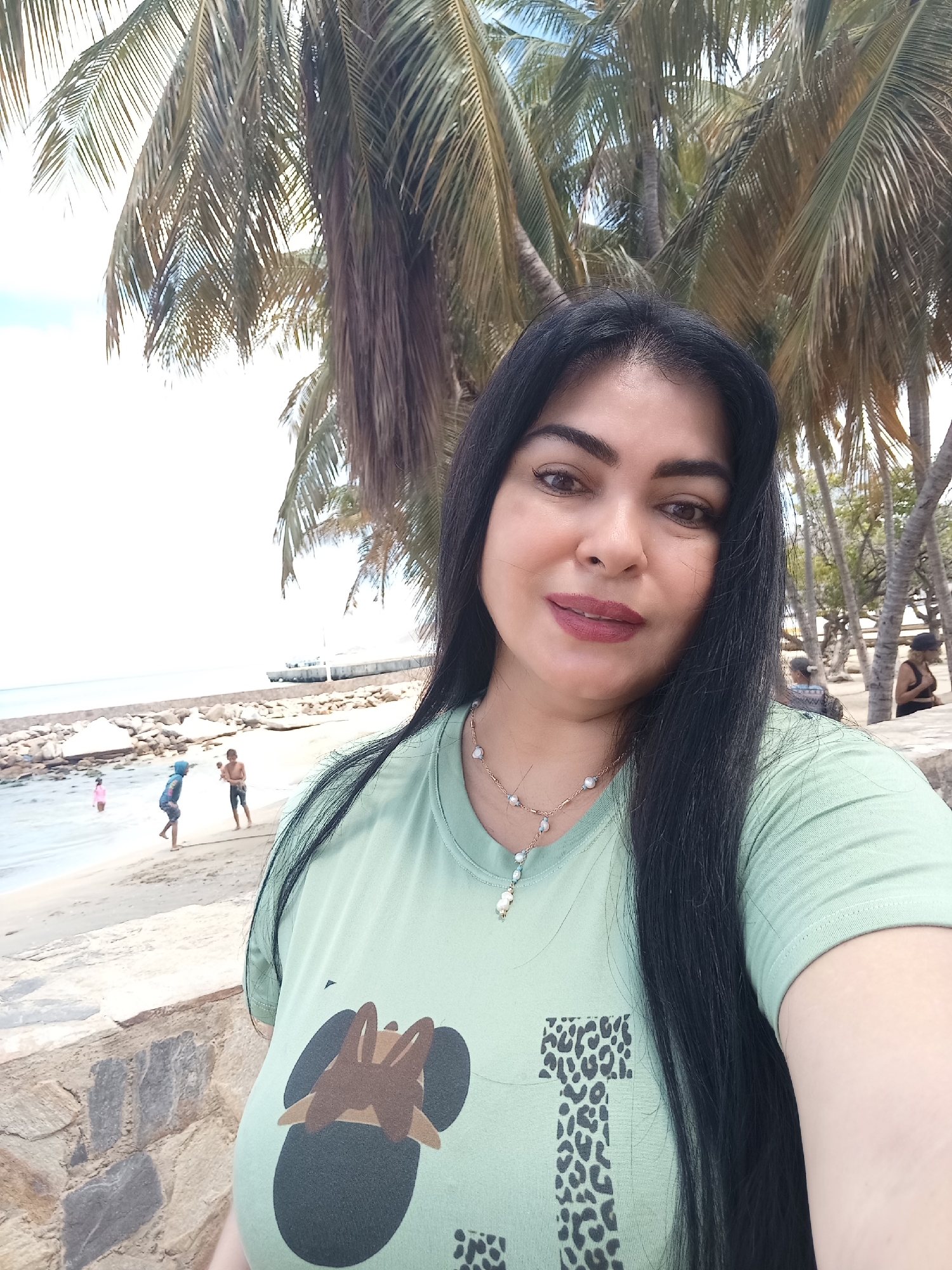 The image shows a woman taking a selfie on a beach in Juan Griego, Venezuela. The woman has long, black hair and is wearing a light green t-shirt with an animal print design. She is also wearing a necklace.
In the background, there is a sandy beach with several people, including children playing in the water. A stone wall separates the beach from the area where the woman is standing. Palm trees provide shade. A pier or breakwater extends into the ocean in the distance.
