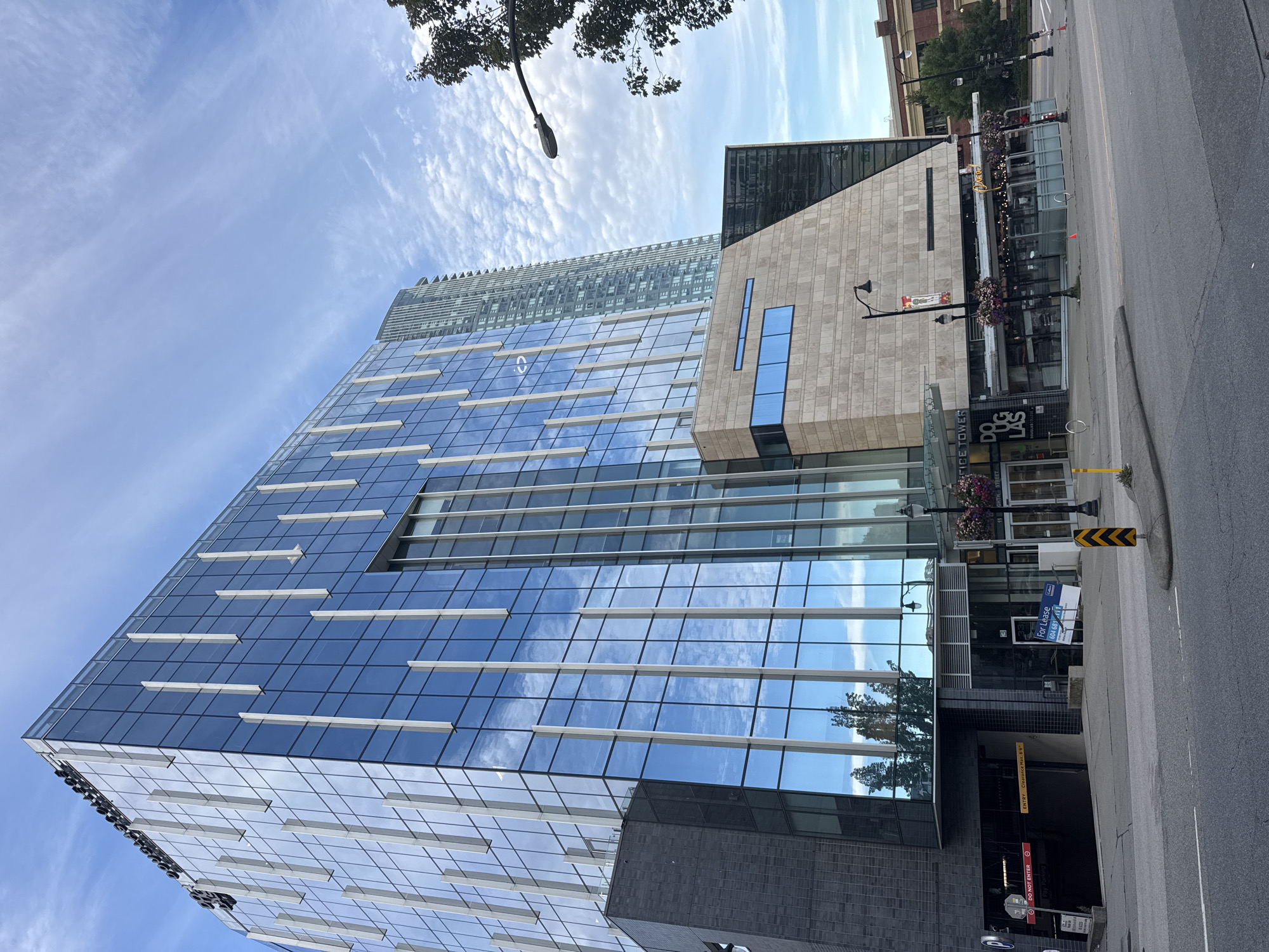 This is a photograph of the Douglas Centre, a modern mixed-use building located at 700 Douglas Street in New Westminster, Canada. The building features a prominent glass facade with vertical white elements and a lighter stone-clad section. In the foreground, a street with two lanes of traffic runs parallel to the building. A parking garage entrance is visible on the left side of the image, marked with red and white signage. On the right side of the building's ground floor, there is a storefront with outdoor seating and hanging flower baskets. A lamppost with a banner is situated in front of the storefront. In the background, a taller, more slender residential tower is visible behind the Douglas Centre. The sky is partly cloudy with a blue hue. No people are clearly identifiable in the scene.