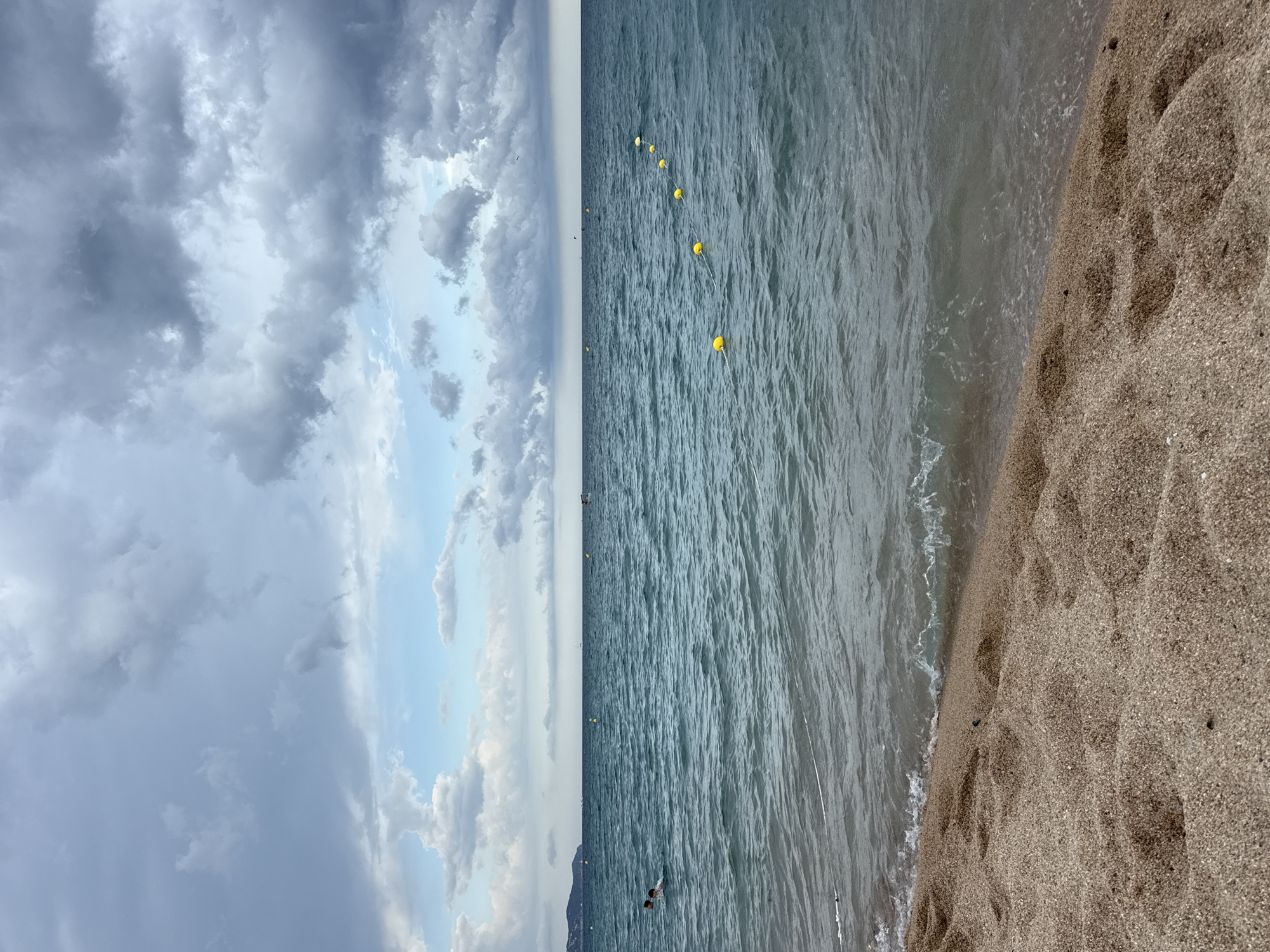 The image depicts a view of the sea and a sandy beach in Blanes, Spain. The foreground features a close-up of the textured sand, with small pebbles and footprints visible. Gentle waves are breaking on the shore. In the middle ground, the turquoise sea stretches out, with a line of yellow buoys marking a boundary in the water. Two people are swimming in the sea to the left of the frame. The background shows the horizon with a distant coastline and a few boats. The sky is filled with dramatic, dark grey clouds interspersed with patches of lighter blue sky.