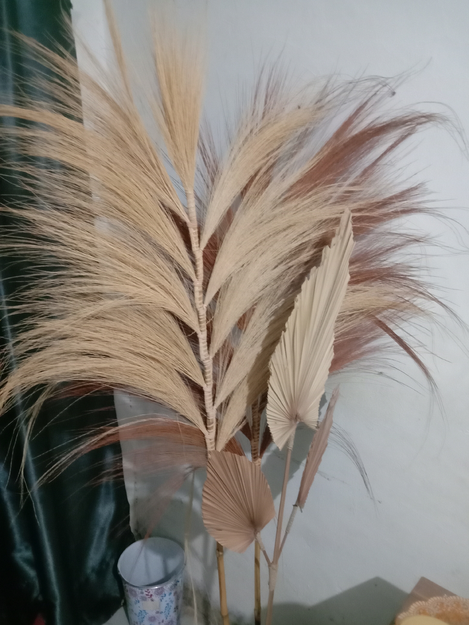The image shows a close-up of dried decorative plants arranged in a vase, likely indoors. The plants are predominantly light brown and beige with some reddish-brown hues, featuring feathery fronds and fan-shaped leaves. They are displayed against a light blue-grey wall on the right and a dark green, silky fabric on the left. A small, patterned cylindrical object, possibly a can or a small vase, is visible near the bottom left corner. The lighting appears to be soft, casting subtle shadows. The scene suggests a home or interior decoration setting. Without further information or context, it is impossible to ascertain the specific location within Kwandang, Indonesia.