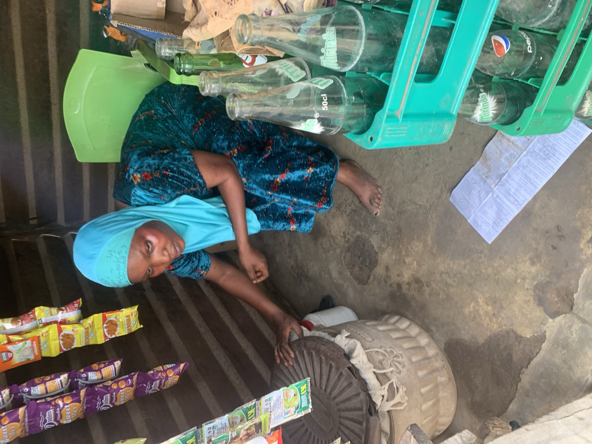In Shagamu, Nigeria, a woman wearing a teal headscarf and a floral print dress is seen tending to a stall. She is crouched down, with her left hand resting on a large, textured circular object, possibly a millstone, positioned atop a woven container. Her right hand is extended, and she appears to be reaching for something on the ground.
The stall is stocked with various items, including rows of colorful snack packets hanging from above. To the right of the woman, several green plastic crates are filled with glass bottles, some of which are labeled "Mirinda" and "Pepsi." A green plastic chair is visible behind her. The ground beneath her feet is concrete and appears wet in places, suggesting a recent spill or cleaning. A folded piece of paper lies on the ground near the bottles. The background consists of slatted wooden walls, indicating an outdoor or semi-enclosed market setting.