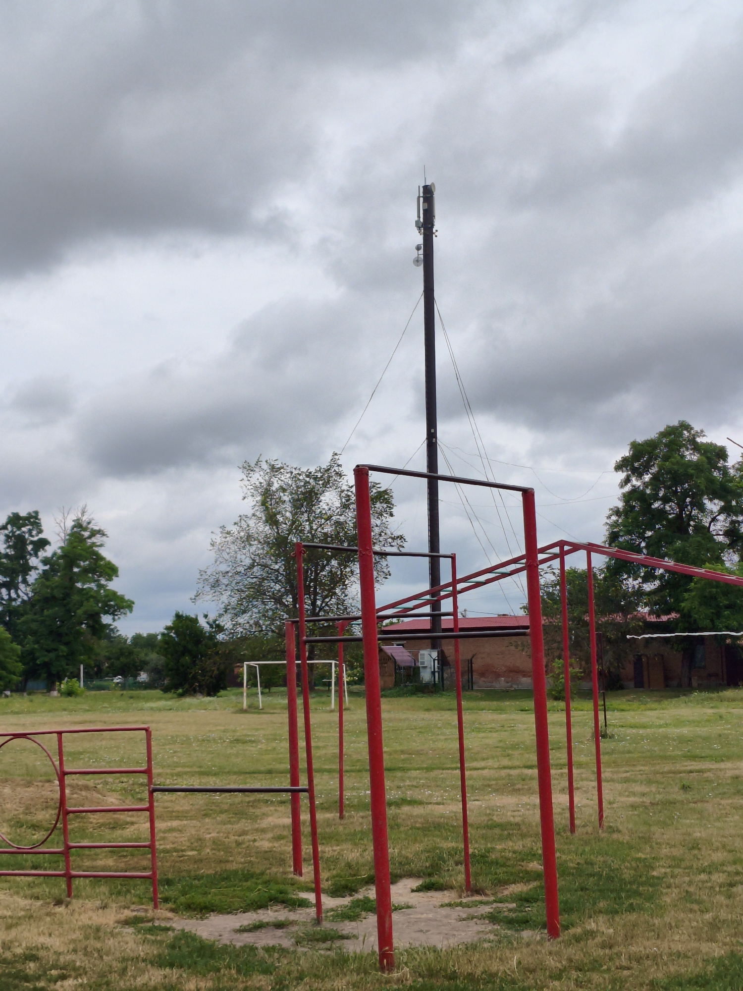 The image depicts an outdoor recreational area with various exercise equipment. In the foreground, there is a set of red metal bars, likely part of a pull-up or monkey bar structure, set on a grassy field. To the left, another red metal frame with horizontal bars and a circular element is visible. In the background, trees and a brick building can be seen. Further back, a white soccer goal is positioned on the grass, suggesting the area is a sports field or park. Dominating the upper portion of the image is a tall, dark pole with antennas and equipment, resembling a telecommunications tower. The sky is overcast with heavy grey clouds. The scene appears to be located in the city of Ustyvytsia, Ukraine, given the provided context. There are no people or animals visible in the image.