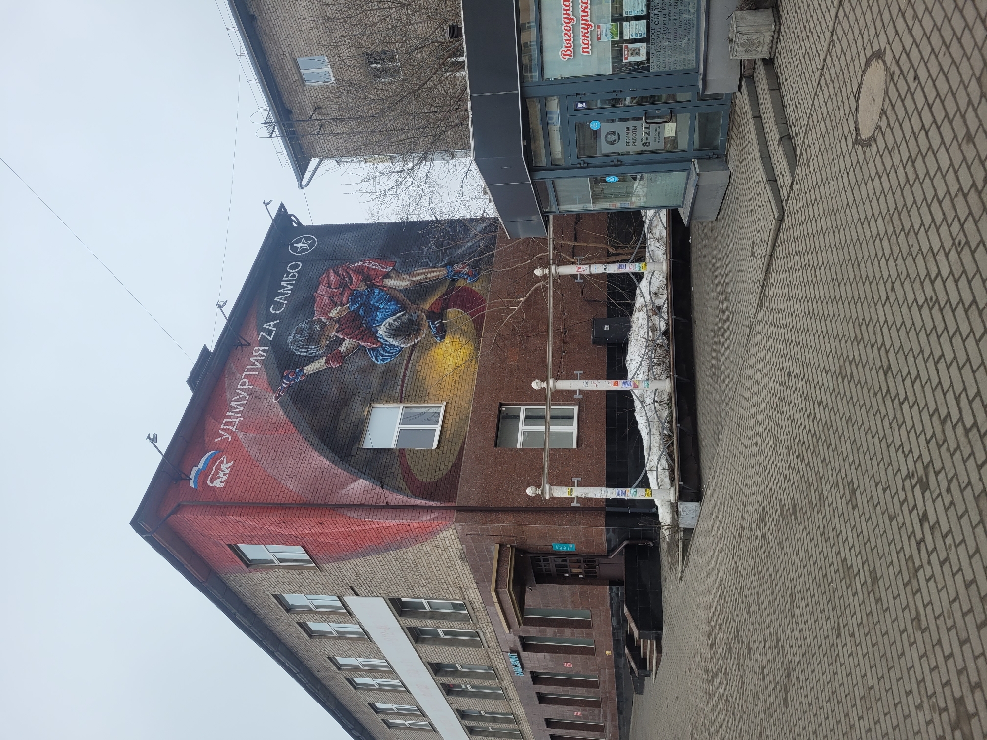 Here is a detailed description of the image:
**Scene:** An urban view in Izhevsk, Russia.
**Subject:** The focal point is a multi-story brick building, where a mural dominates one of its side walls.
**Mural:** The mural portrays two Sambo wrestlers engaged in a grapple. The text "Удмуртия за Самбо" (Udmurtia for Sambo) is written in Cyrillic script along the top of the mural. The regional flag of Udmurtia is partially displayed on the corner of the building's upper portion.
**Building Exterior:**
* The building has multiple rectangular windows aligned horizontally across its various floors.
* A dark brown extension with signage is attached to the building's side facing the street. The extension looks to be a storefront.
* Power lines run along the facade of the building.
**Storefront:** The storefront has glass windows and doors. A sign indicates opening hours: 8-21 (8 AM to 9 PM).
Another sign, written in Cyrillic, promotes products like yogurt, cheese, and other dairy products.
**Streetscape:**
* The foreground shows a paved sidewalk, constructed with interlocking rectangular bricks.
* Miscellaneous poles and signage are along the sidewalk.
**Environment:** The sky appears overcast and gray. Bare tree branches are visible.