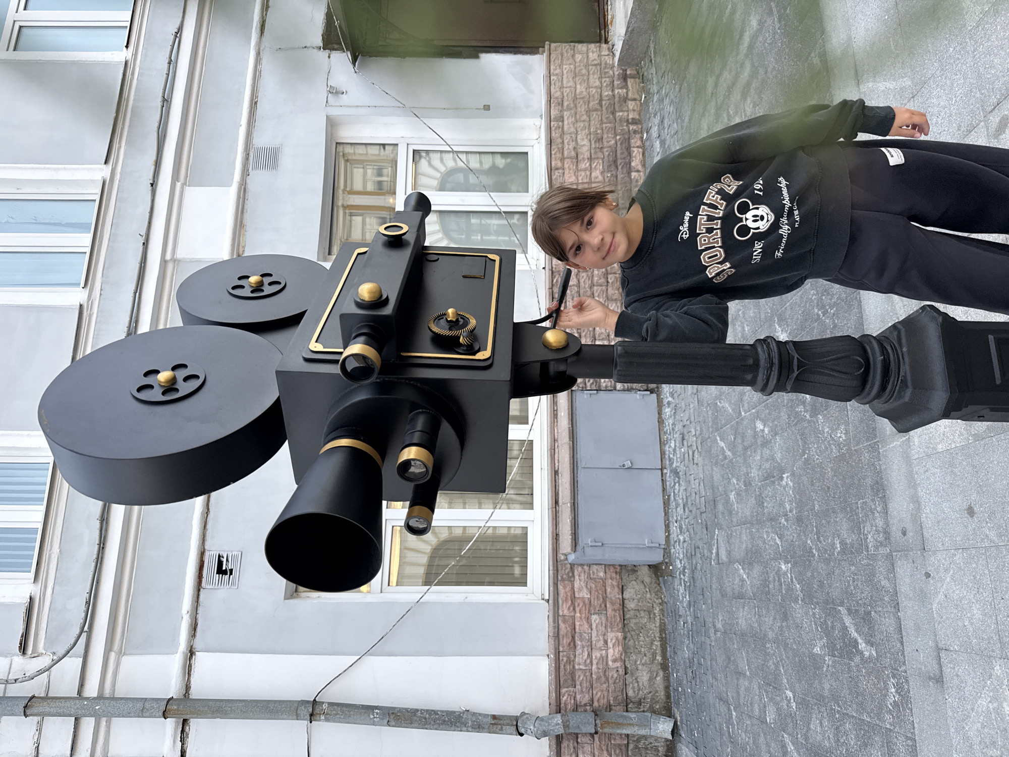A young girl with brown hair is standing next to a large, black, decorative film camera sculpture on a lamppost. The sculpture is detailed and appears to be made of metal. The girl is wearing a dark gray sweatshirt with a Disney Sportif graphic and black sweatpants. She is looking at the camera and touching the side of the sculpture. The scene takes place on a cobblestone street in front of a building with a light gray facade, brown brick accents, and white-framed windows. The setting is likely an outdoor urban environment.