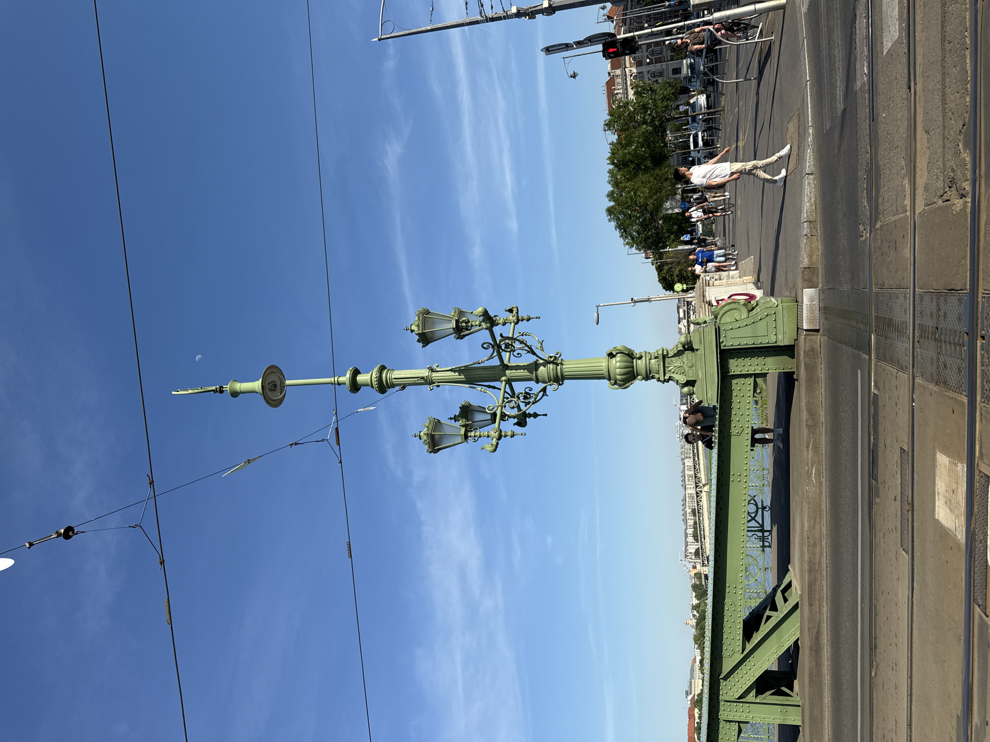 The image captures a daytime scene on the Széchenyi Chain Bridge in Budapest, Hungary. The photograph features a green, ornate lamppost in the foreground against a clear blue sky with thin clouds. Electrical wires stretch across the sky to the left of the lamppost.
In the background, people are walking and cycling across the bridge. Buildings can be seen in the far distance on the Buda side of the Danube River. A traffic light is visible in the background. The bridge's structure, including the road surface and the metal framework, is also visible.