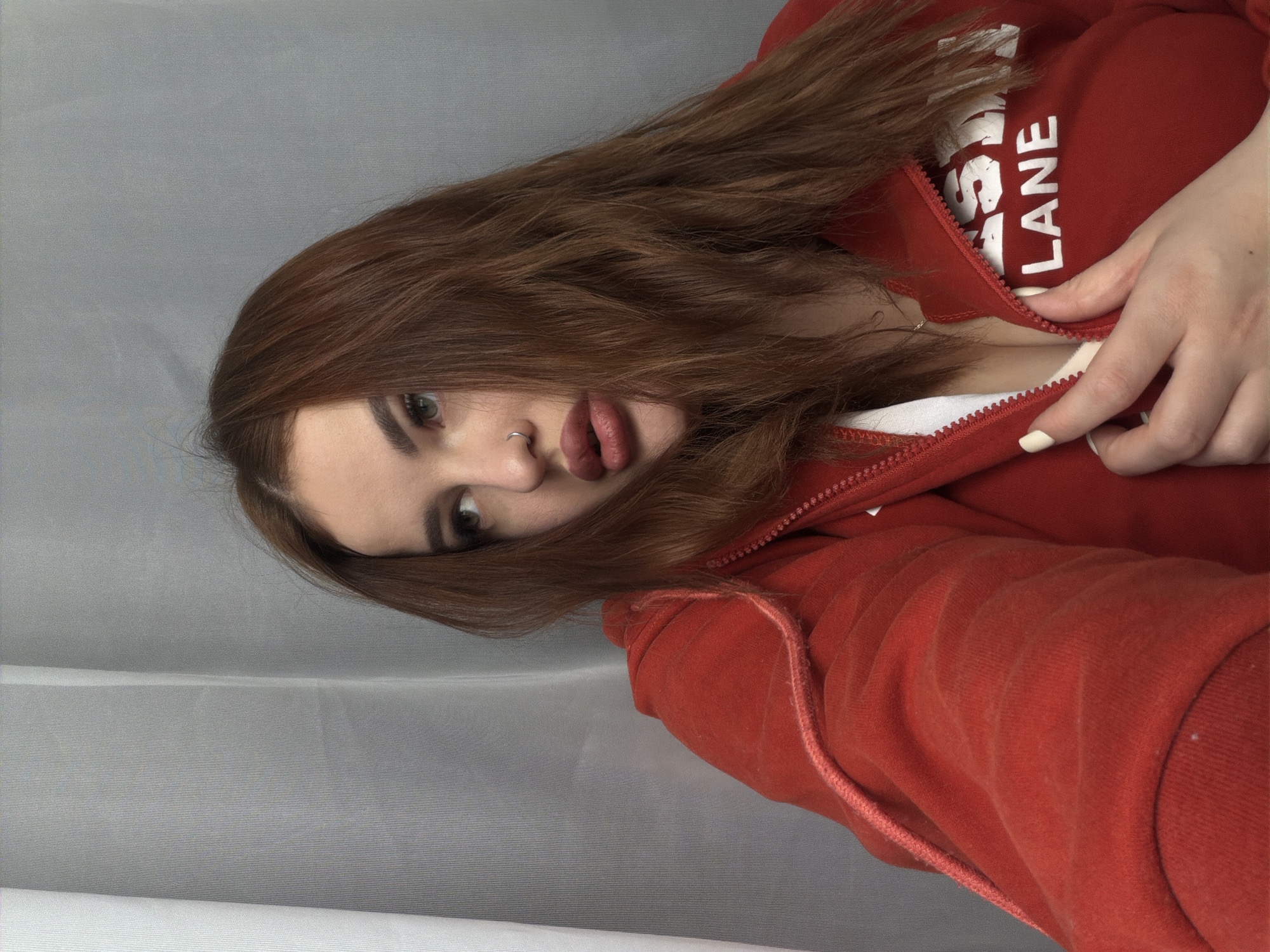 A woman with long brown hair, wearing a red zip-up hoodie, is posing for a selfie. Part of her face is obscured by her hair. Her lips are accentuated with a dark lipstick, and she is wearing a nose ring. She is partly unzipping her hoodie. The background is a gray fabric. There is no verifiable information about the location of this image.