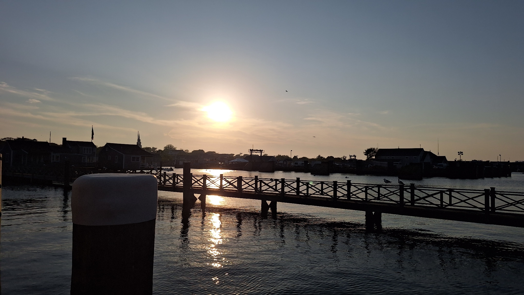 The image shows a harbor scene in Nantucket, Massachusetts, USA, during sunset. The sun, a bright orb, is positioned in the upper left quadrant of the sky, casting a warm glow and reflecting on the water. A wooden bridge with crisscrossed railings spans across the water, leading to a silhouetted shoreline with several buildings and a church steeple. The water is calm, with ripples reflecting the sunlight and the bridge. A large, white, cylindrical object, likely a buoy, is prominently in the foreground on the left. Seagulls are visible on the bridge and near the water. The overall atmosphere is serene and tranquil, typical of a coastal town at dusk.