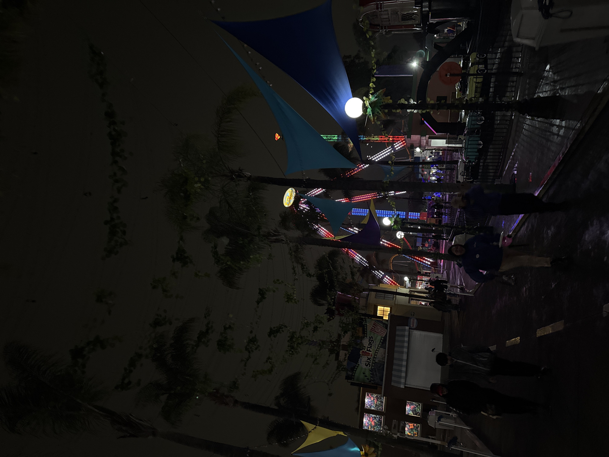 At night, the Six Flags Mexico theme park in Mexico City is illuminated with colorful lights, and several people are seen walking. A Ferris wheel with lights is in the background, as well as other rides with colorful lights. There are trees and shade sails, and a building with a "Six Flags" advertisement.
