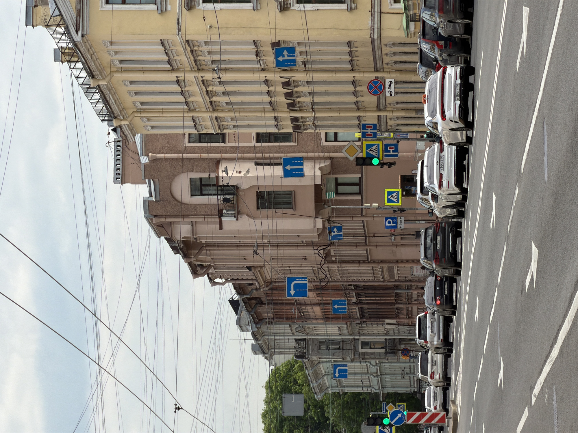 This image captures a vibrant street scene in an urban environment, likely in Saint Petersburg, Russia, based on architectural styles and visible cues. The perspective is from the street level, looking along a row of buildings.
The scene features several buildings of varying architectural styles, some with ornate facades in light yellow and beige hues, others in more subdued browns and grays. Many windows are visible, and some buildings have balconies with metal railings. A dense network of overhead wires crisscrosses the sky, suggesting trolleybus lines or extensive utility infrastructure.
Along the right side of the street, a row of cars is parked, facing away from the viewer. These include several white sedans (some with taxi lights on their roofs) and darker SUVs. The street itself is paved with asphalt and features distinct white lane markings, including arrows indicating directions. A prominent traffic light displays a green signal.
Numerous road signs are visible, providing navigation and regulatory information. These include blue square signs with white arrows indicating mandatory directions (such as "left turn only," "forward or left," "forward only," and "right turn only"). Other signs include a red circular "No Stopping" sign, a yellow diamond "Priority Road" sign, and blue square signs indicating "Pedestrian Crossing." One parking sign displays "P" with "↑ 30m ↑" text, suggesting parking is available 30 meters ahead. A temporary red and white striped barrier-like sign bears the Cyrillic text "СТОЙ" (STOJ - STOP). A small sign with a tow truck icon indicates a tow-away zone.
The weather appears to be overcast but bright, characteristic of daytime. License plates on the parked cars are visible, including "H384EE 799", "T340AO 199", and "P723OH 98". The "98" region code on one plate confirms the location as Saint Petersburg. A lush green tree is partially visible towards the bottom left.