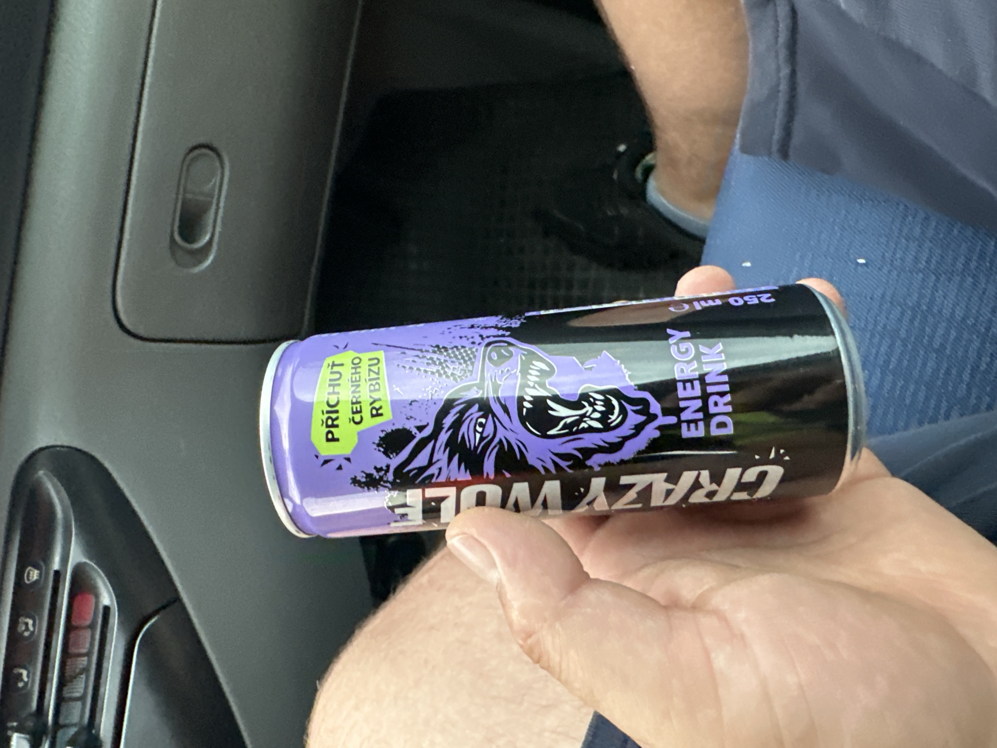 A person holds a can of "Crazy Wolf" energy drink, displaying a purple and black design with a wolf's head graphic. The can is oriented horizontally, with text visible on its surface. Text on the can includes "PŘÍCHUŤ ČERNÉHO RYBÍZU" (Flavor Blackcurrant) in a green box and "ENERGY DRINK" vertically oriented. The brand name "CRAZY WOLF" is also prominently displayed, though parts of it are obscured by the hand holding the can. The background shows a car interior, with a dark gray dashboard and a blue fabric seat. A person's legs are visible in the background, wearing dark shoes. The scene appears to be taking place indoors, likely in a vehicle, given the dashboard and seat. There are no strong indicators of time of day or weather conditions.