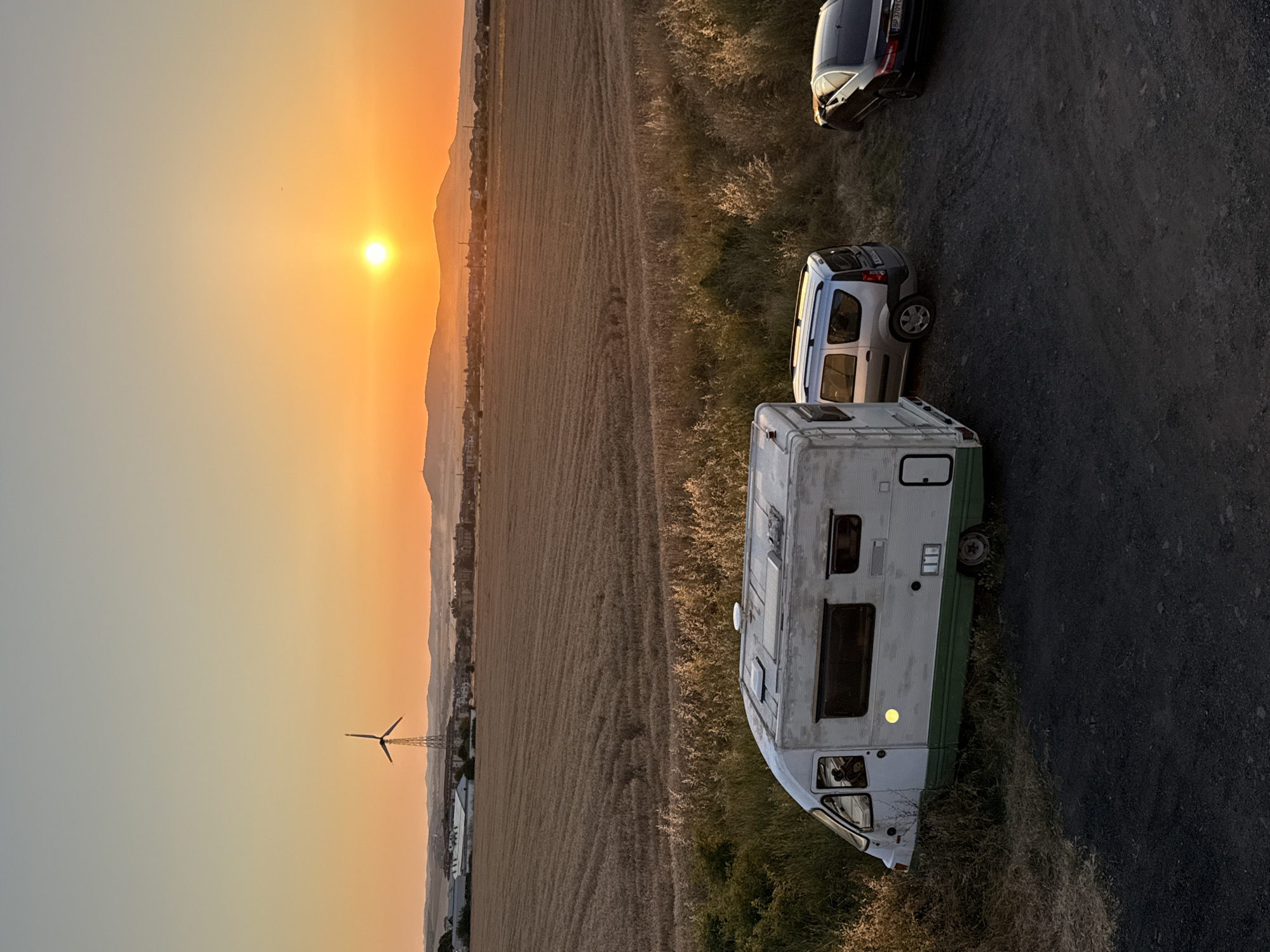 The image shows a rural landscape at sunset, with a golden sky and silhouetted mountains in the distance. The scene features three vehicles parked on a dark, gravelly surface next to a field of dry, brown vegetation. The vehicles include a white and green caravan in the foreground, a silver van behind it, and a dark-colored car further back. The caravan appears to be older, with some visible wear and tear.
A wind turbine stands in the middle ground, to the left of the vehicles, its blades silhouetted against the bright sky. Beyond the field, a small town or village is visible, with buildings clustered along the foothills. The overall atmosphere is serene and peaceful, suggesting a quiet moment of rest or a stop during a journey. The time of day is clearly sunset, indicated by the warm, orange hues of the sky and the long shadows cast by the objects. The location is identified as Ravda, Bulgaria. No people are visible in the image. There is some text visible on the license plate of the dark-colored car, but it is too blurry to read.