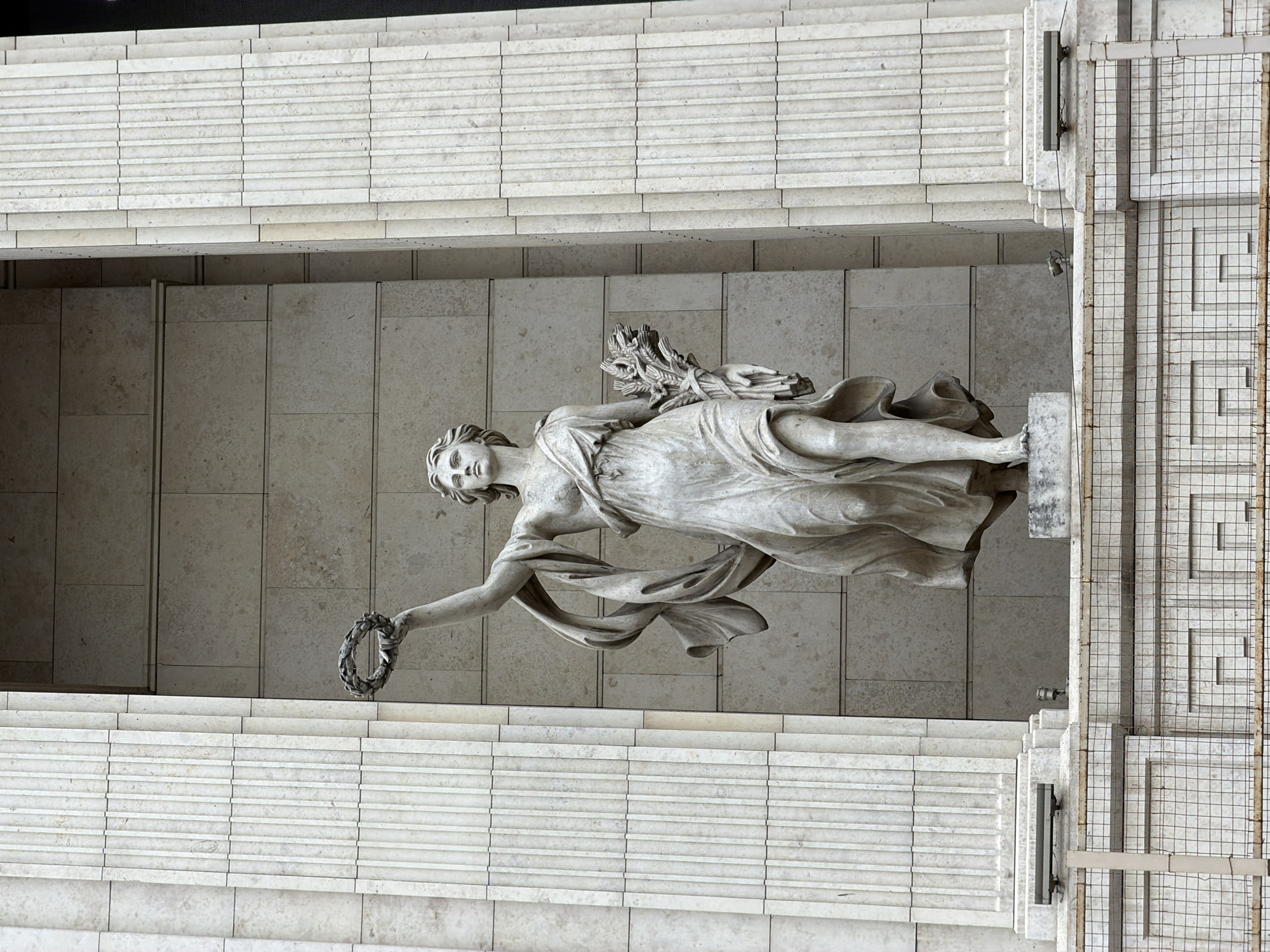 The image is a slightly angled overhead shot of a statue of a draped female figure holding a wreath in her extended left hand. She rests on a plinth made of the same pale stone as the surrounding architecture. She holds a bundle of sheaves in her right arm. The statue is surrounded by a multi-layered structure of horizontally stacked pale-colored stone, suggestive of an architrave or a framed space. To the right of the statue, a section of the architecture features a lattice-like grill. The pale, neutral color palette suggests the use of marble or limestone.
Based on the architecture and style of the statue, this image is likely of the statue of "Glory" on the General Staff Building in Saint Petersburg, Russia.
