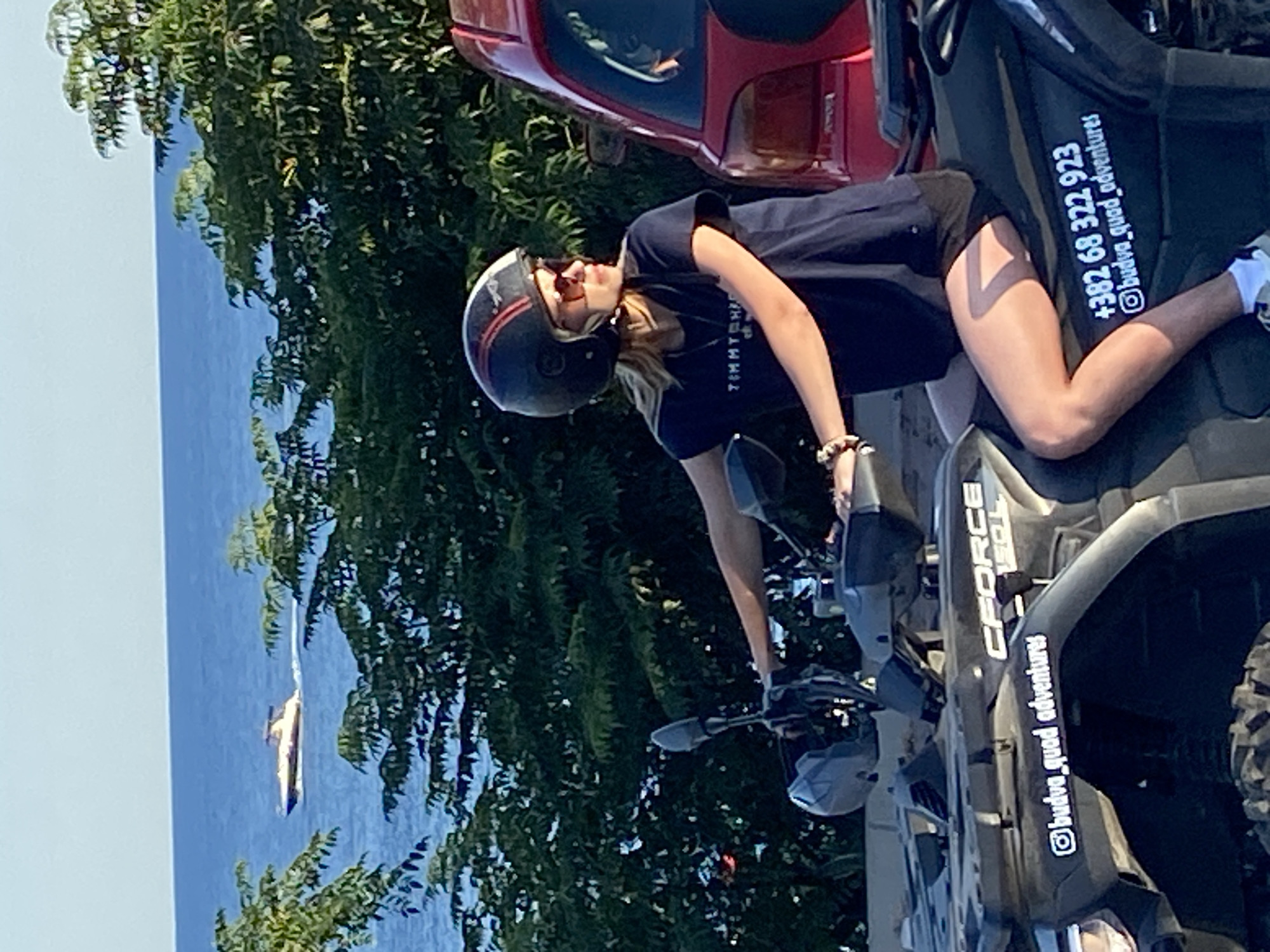 In Prijevor, Montenegro, a woman wearing a helmet and sunglasses is seated on an all-terrain vehicle (ATV). She is looking to her right. The ATV has the words "CFORCE" and "budva_quad_adventures" on its side, along with a phone number "+382 68 322 923". She is wearing a dark blue t-shirt and black shorts, with white sneakers. Behind her, a red vehicle is partially visible. The background shows green trees and the blue sea with a boat on the water. The scene appears to be outdoors, possibly near a tourist destination.