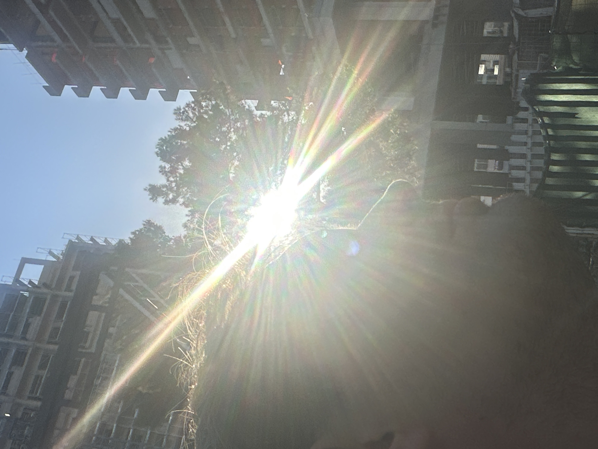 A low-angle shot directed upwards, heavily overexposed by the sun, reveals a fragmented view of Budva, Montenegro. Architectural elements, including buildings and construction scaffolding, are visible against a bright blue sky. The silhouette of a person is partially visible in the foreground, obscuring some of the scene. The strong sunlight creates lens flare and makes it difficult to discern finer details.