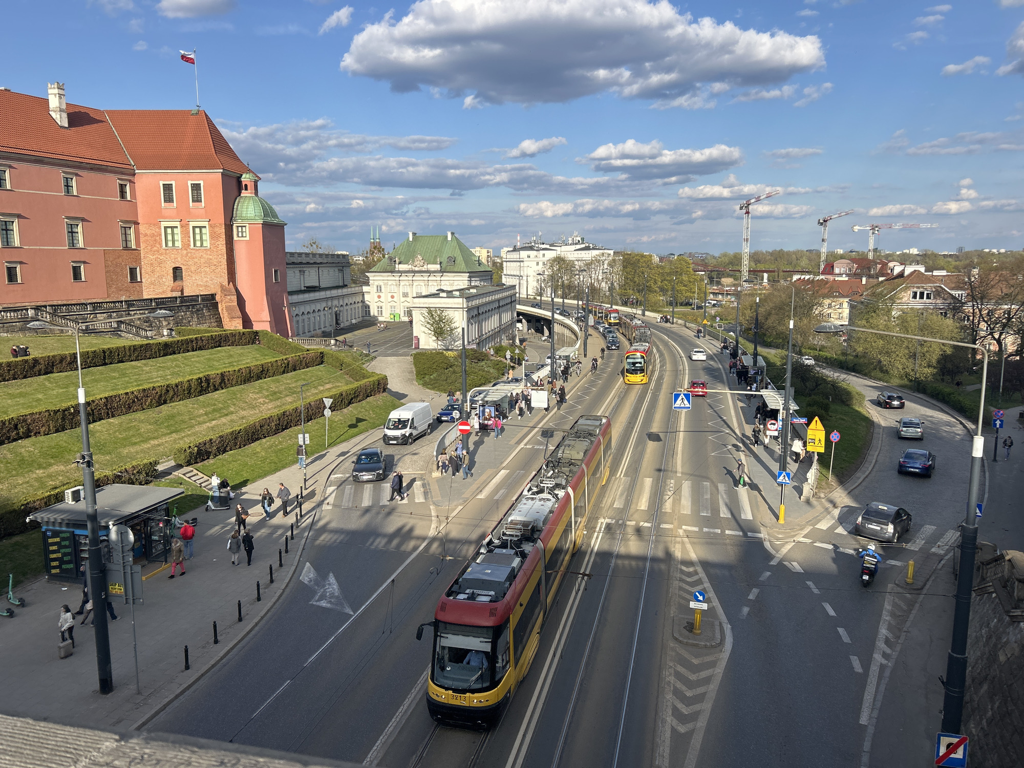 This is an elevated view of Warsaw, Poland, showing a busy street scene. In the foreground, a yellow and red tram is traveling along the tracks in the middle of the road. Pedestrians are crossing at the designated crosswalks, and several cars and a scooter are navigating the road. A newsstand stands on the left side of the road. The Royal Castle of Warsaw is visible on the left side of the frame. In the background, there is a mixture of older and modern buildings, trees, and a bridge under construction with cranes. The sky is bright blue with scattered clouds.