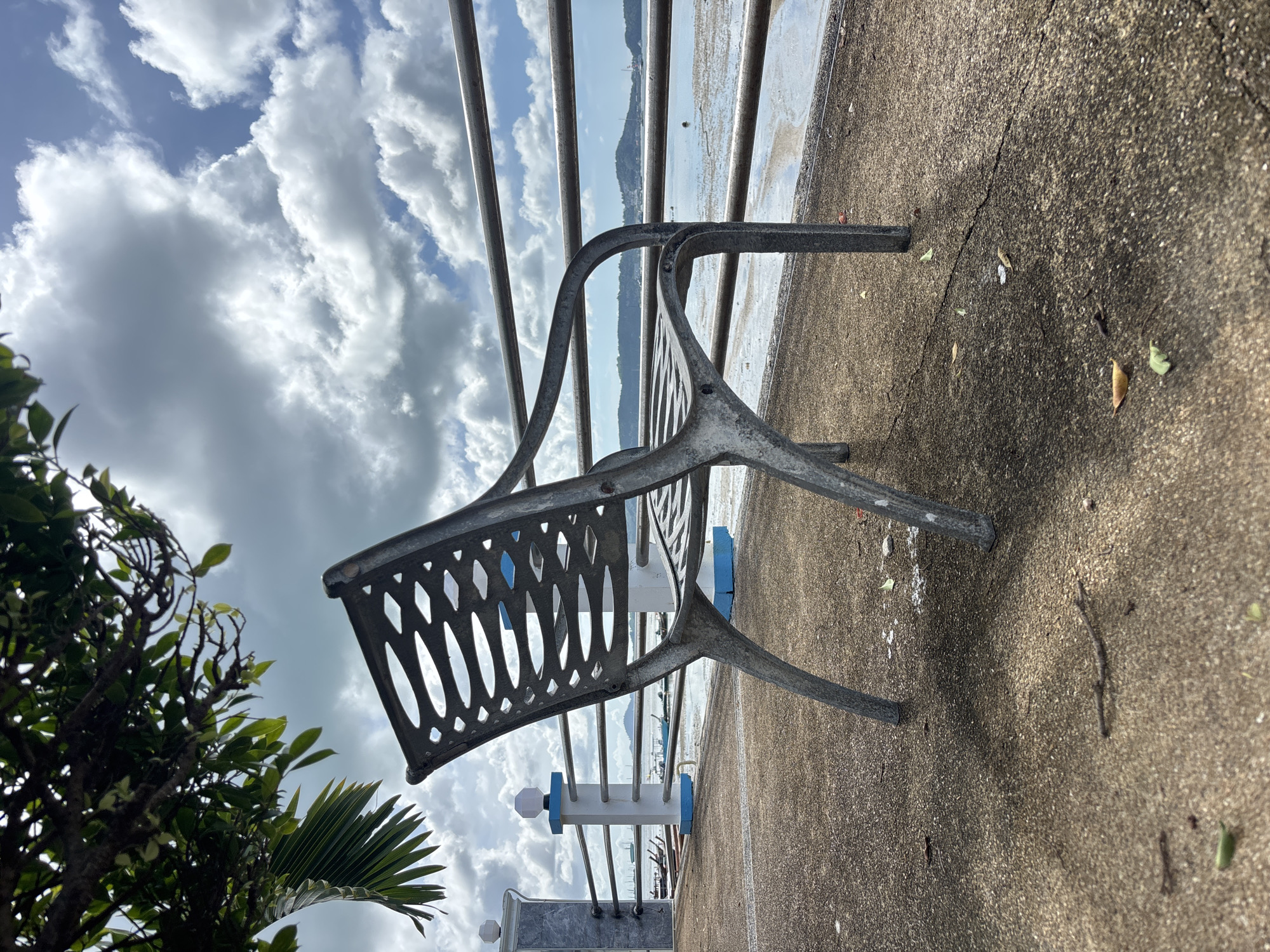 A weathered metal bench with a patterned seat and backrest sits on a concrete walkway in Rawai, Thailand. The bench is angled towards the right, facing a body of water. In the background, a metal railing runs horizontally along the walkway, separating it from the water. Beyond the railing, a calm sea is visible with several long-tail boats moored or anchored. The water's edge shows exposed sand or mud flats, indicating a low tide. A distant, tree-covered hill or island forms the horizon. The sky above is partly cloudy with a mix of bright blue patches and large, white cumulus clouds. To the left of the bench, a decorative white pillar with blue accents stands near the railing, and tree branches with green leaves overhang the scene from the upper left. The concrete walkway appears textured and shows some scattered leaves and debris, as well as shadows cast by the bench and foliage.