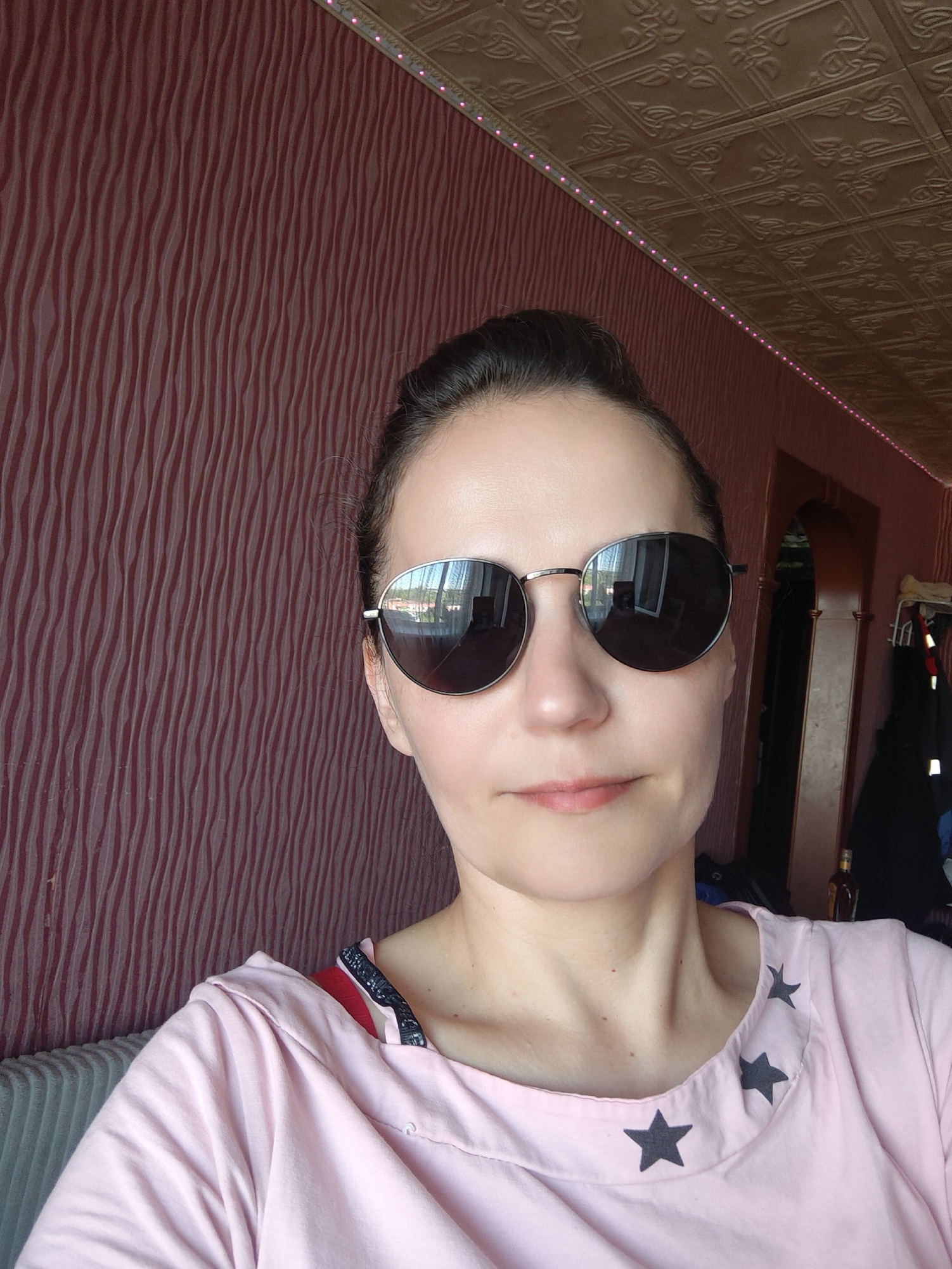 A young woman with dark hair pulled back and wearing round, dark-lensed sunglasses is shown in a selfie. She is wearing a pale pink t-shirt with black star appliqués around the neckline. The background consists of a textured reddish-brown wall and a ceiling with decorative tiles. A doorway is visible on the right, with clothing hanging in the background. The image appears to have been taken indoors, possibly in an apartment in Zheleznogorsk, Russia. The lighting is soft, and the overall impression is casual and relaxed.