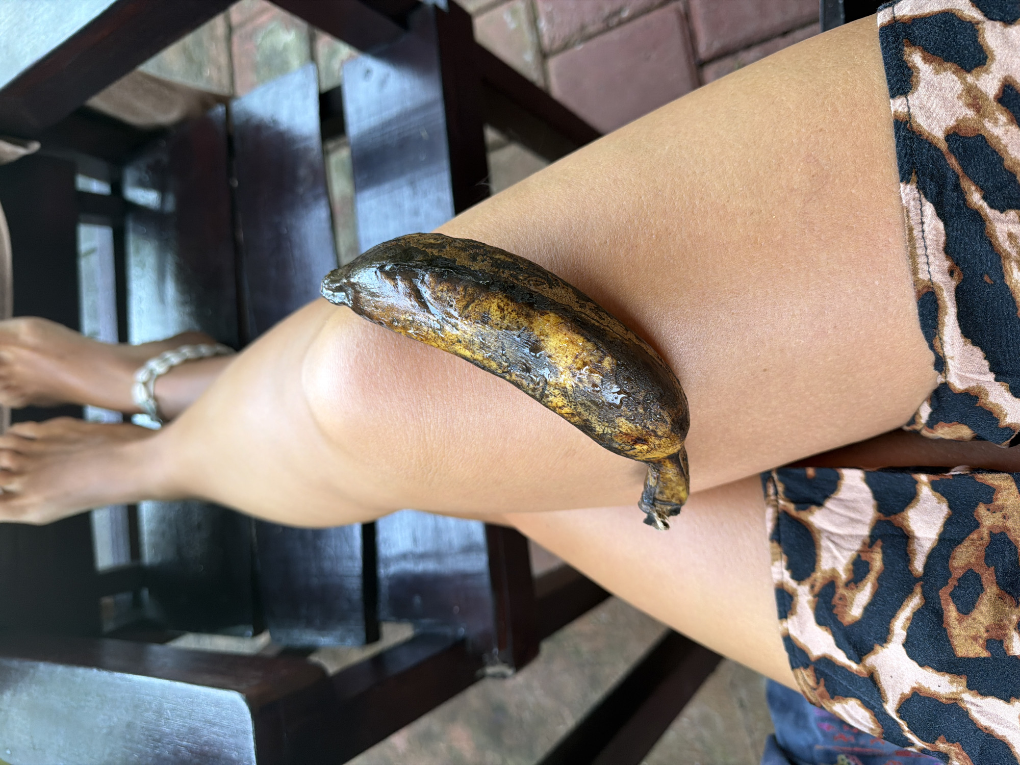 The image presents a close-up view of a person's leg in what appears to be an outdoor setting. The person is seated, as suggested by the presence of a wooden chair on the left side of the frame. A heavily ripened, almost black banana rests on the person's thigh. The individual is wearing a garment with a leopard print pattern. A bracelet is visible around the ankle of the leg extending into the lower-left of the frame. The setting is identified as Kutuh, Indonesia, based on the geo-location data. The flooring appears to be made of bricks.