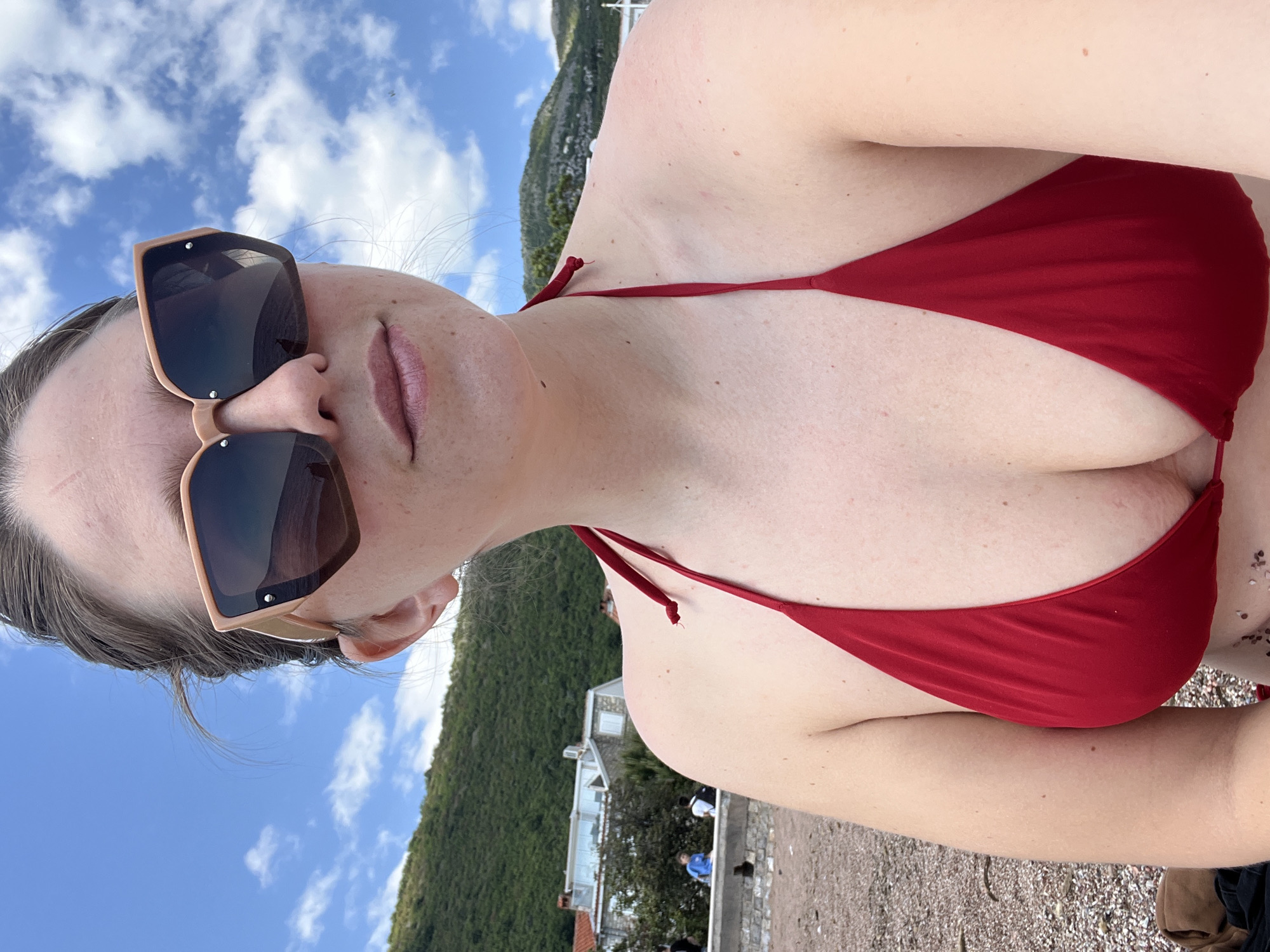 A woman wearing sunglasses and a red bikini top is positioned on a beach. Behind her is a landscape featuring a hillside covered in dense green vegetation. Closer to the beach, there is a building, potentially a residential or commercial structure, with people visible near it. The sky in the background is blue with scattered clouds. The ground beneath the woman appears to be a mix of sand and small pebbles. The location is Petrovac centar, Montenegro.