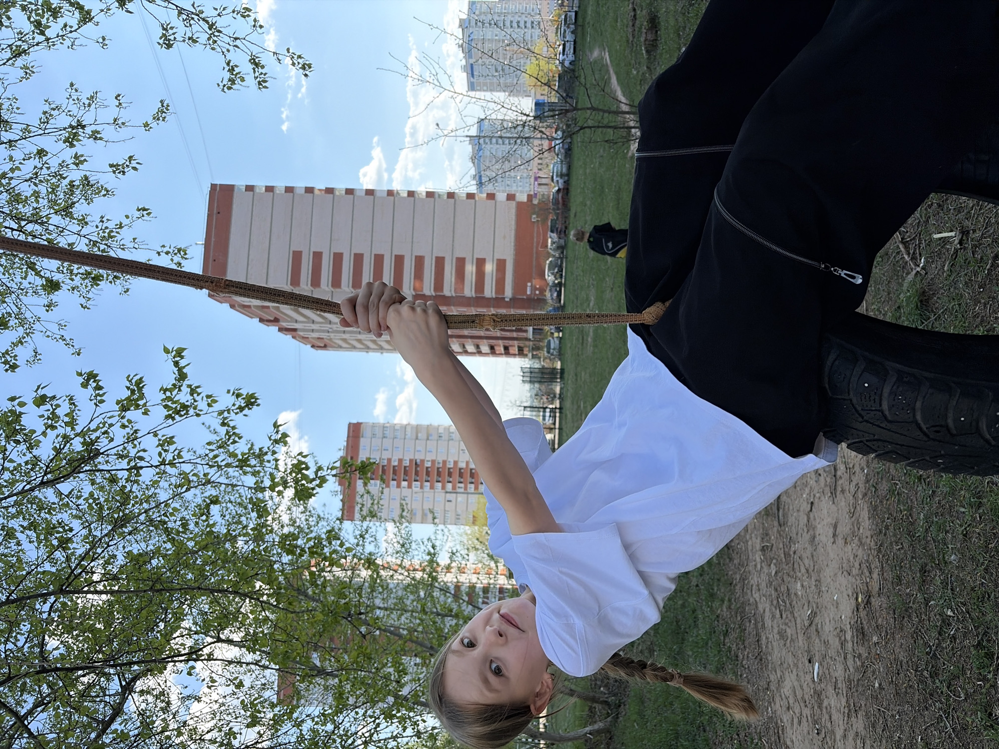 A young girl is swinging on a tire swing in Izhevsk, Russia. She is holding onto a rope attached to the tire. The girl is wearing black pants with zippers and a white t-shirt. She has blonde hair in a braid. In the background, there is a large apartment building with a distinct orange and white striped pattern. The surrounding area appears to be a park with grass and trees. The sky is blue with scattered clouds. In the mid-ground, there is a person who has a black jacket with a yellow stripe and is wearing a black baseball cap.